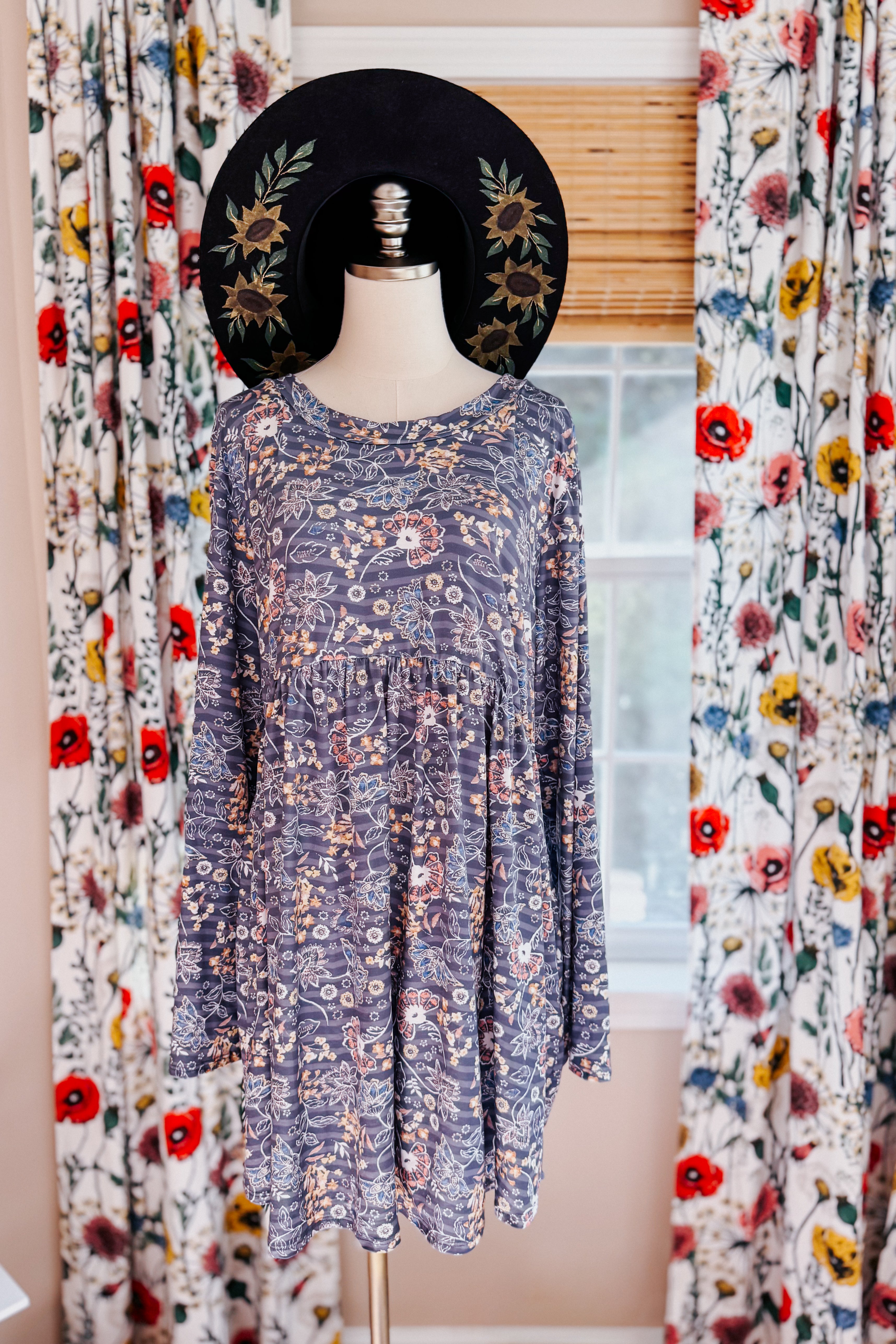 Blue floral patterned swing dress with long sleeves and a gathered waist displayed on a mannequin wearing a black wide-brimmed hat with sunflower embroidery, set indoors near patterned curtains and a window.