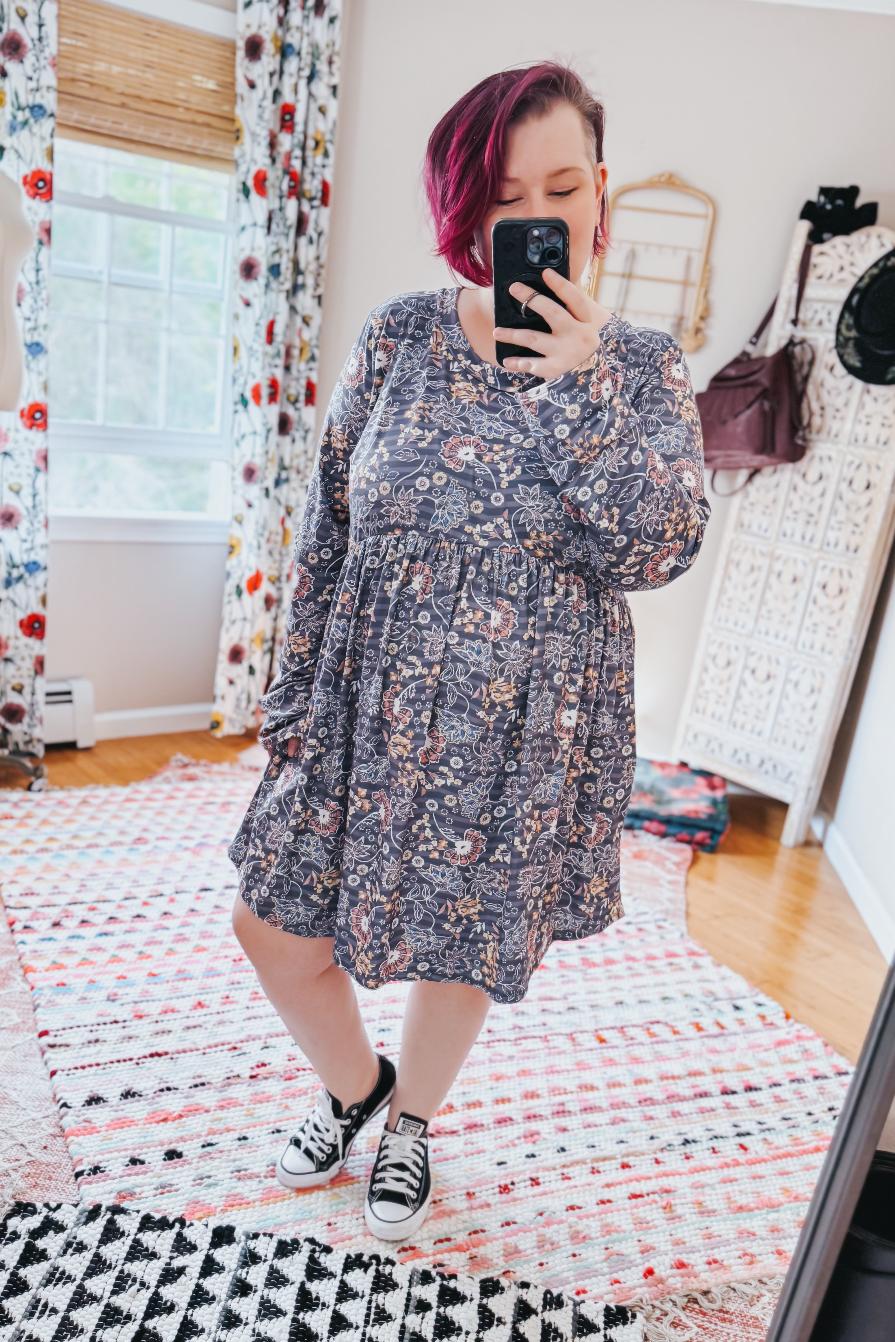 Woman taking a mirror selfie wearing a dark blue floral patterned swing dress with long sleeves and an empire waist, paired with black sneakers in a cozy room.