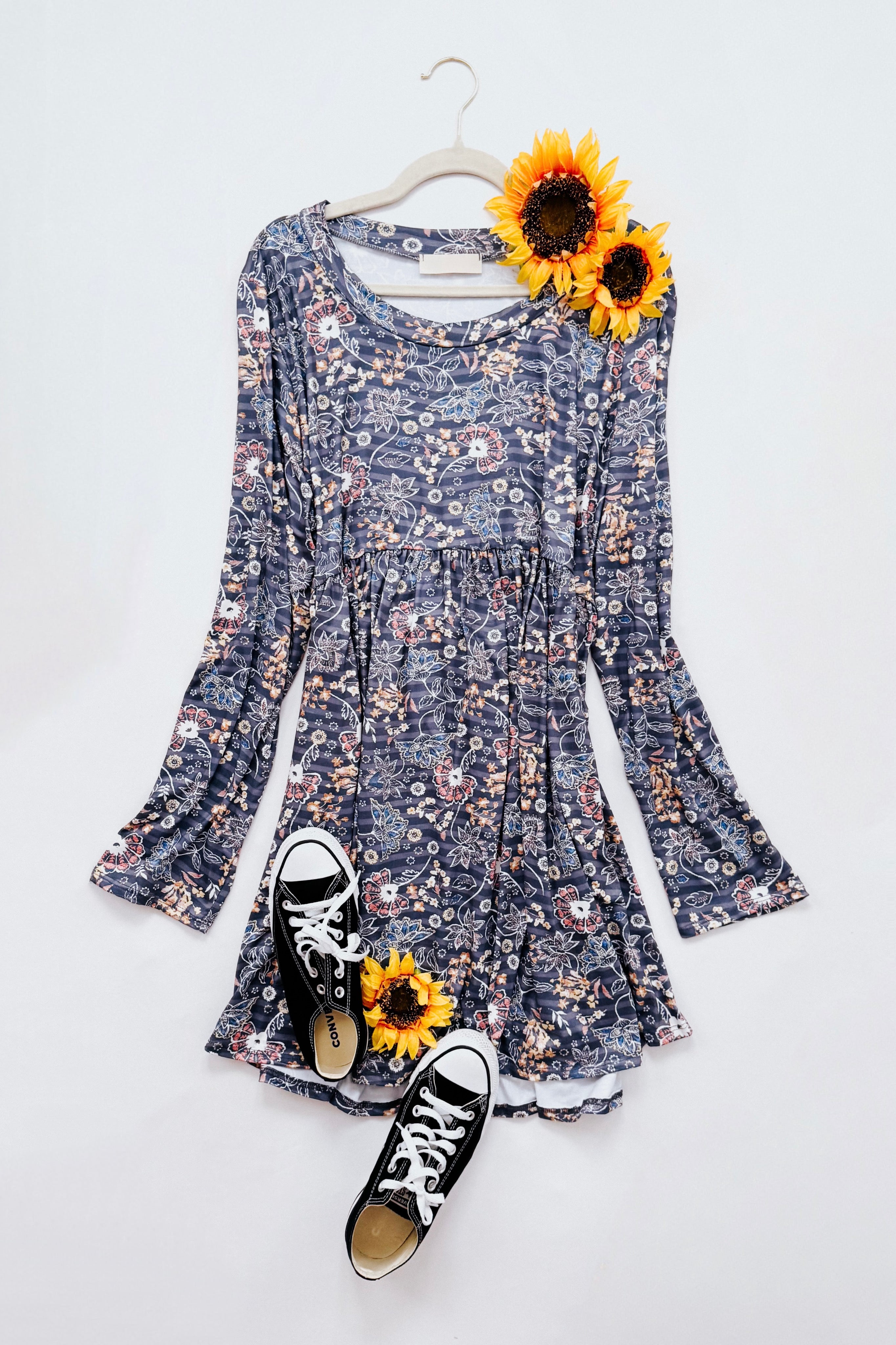 Blue floral print swing dress with long sleeves laid flat alongside black low-top sneakers and two sunflowers on a white background.