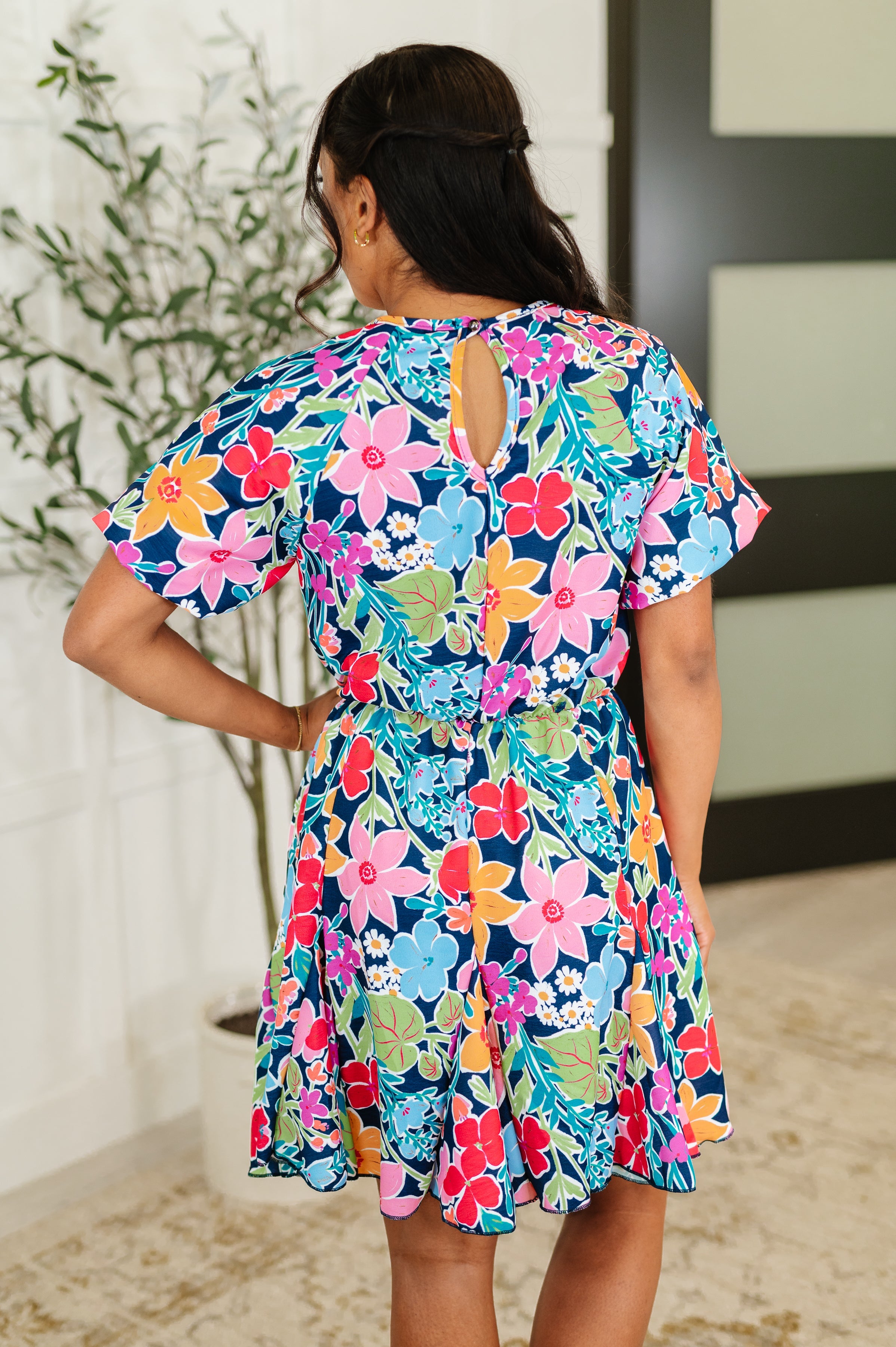 Woman wearing a navy dress with colorful floral print and short flutter sleeves standing indoors with hand on hip