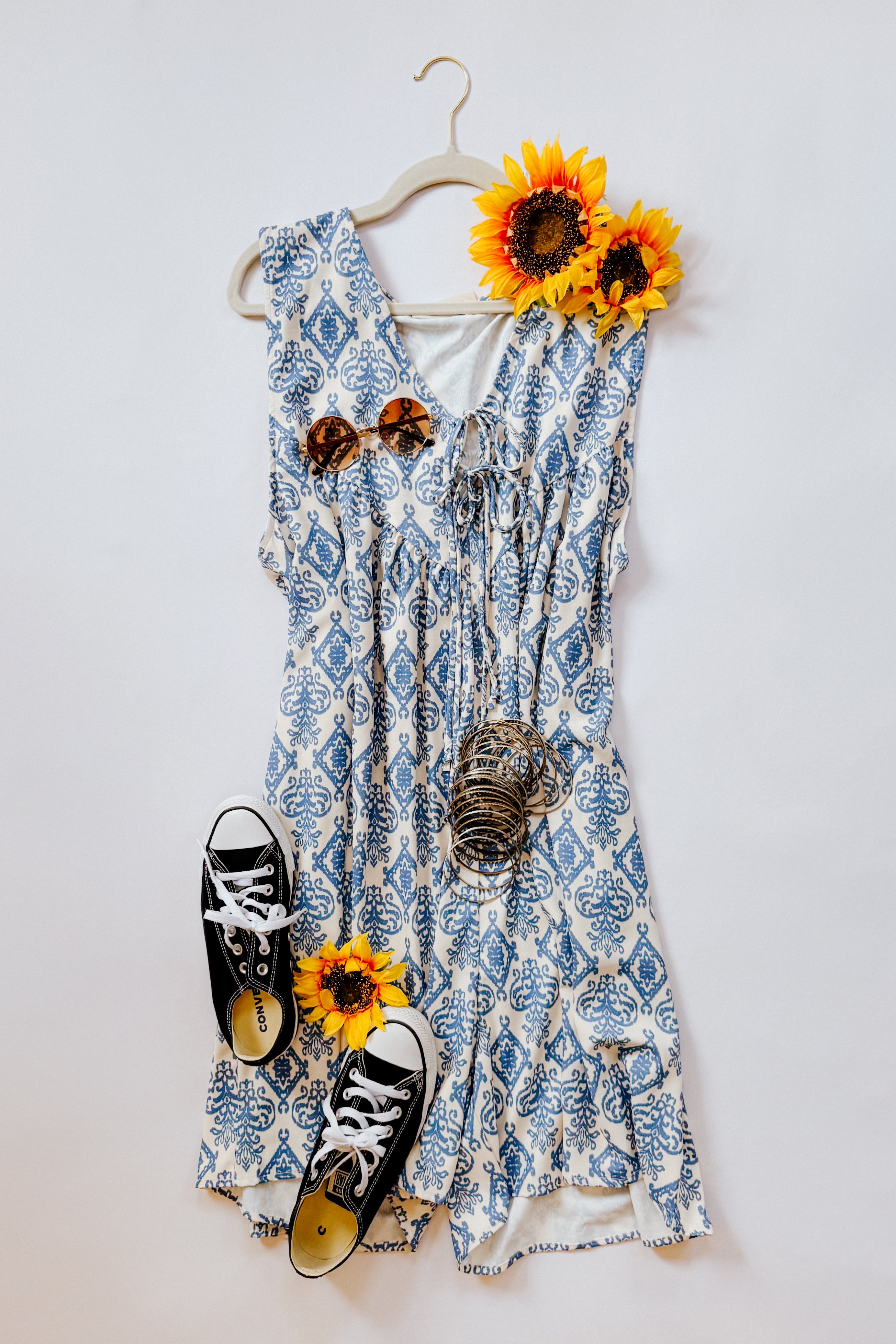Blue and white patterned sleeveless V-neck romper with front tie detail styled with brown sunglasses, stacked bangles, black Converse sneakers, and bright sunflowers