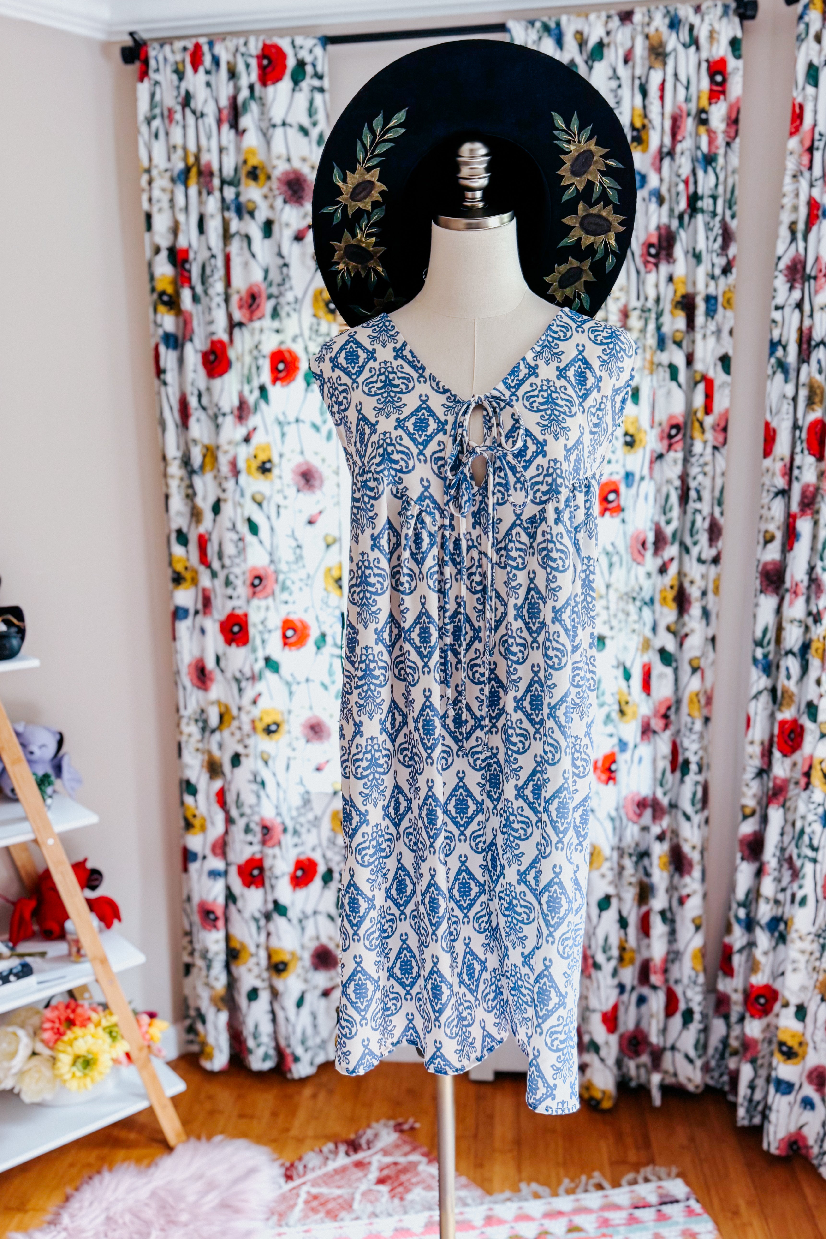 Blue patterned sleeveless V-neck romper with front tie detail displayed on a mannequin in a bright room with floral curtains and a black hat featuring embroidered sunflowers.