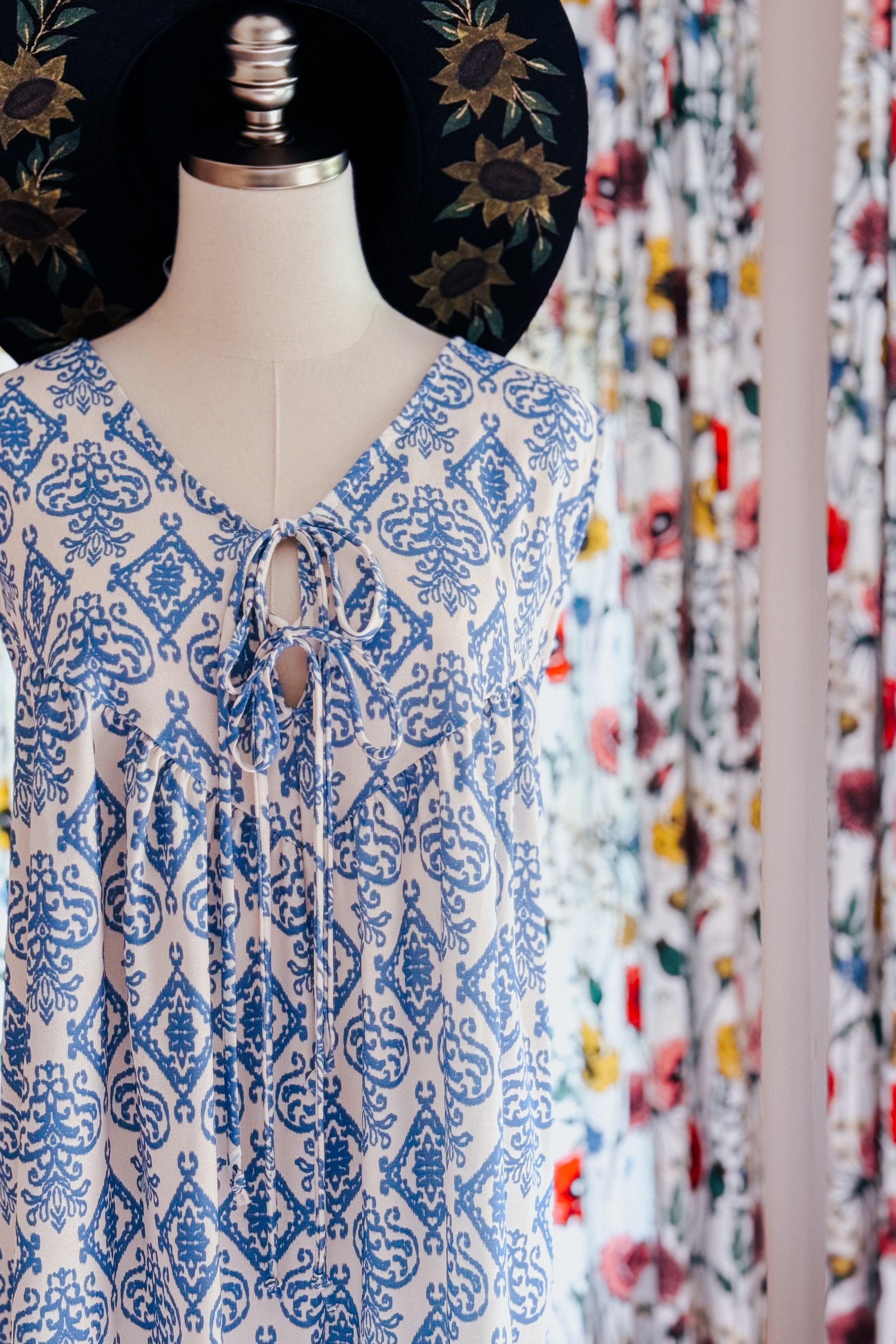 Blue patterned sleeveless romper with a V-neck and front tie detail displayed on a mannequin with a black hat featuring embroidered sunflowers.