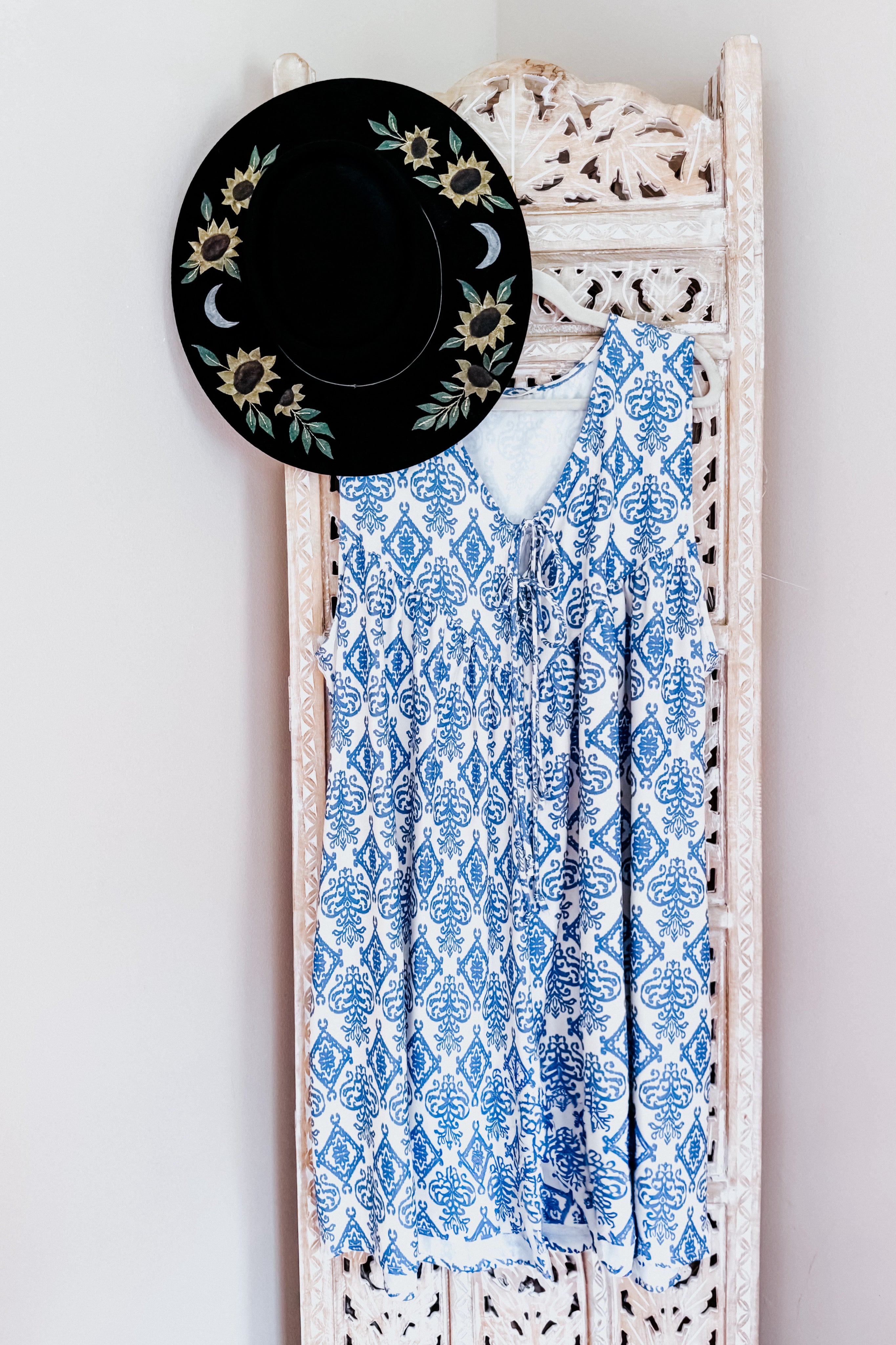Blue patterned sleeveless romper with a V-neck and front tie hanging on a carved wooden screen next to a black hat with sunflower and moon designs