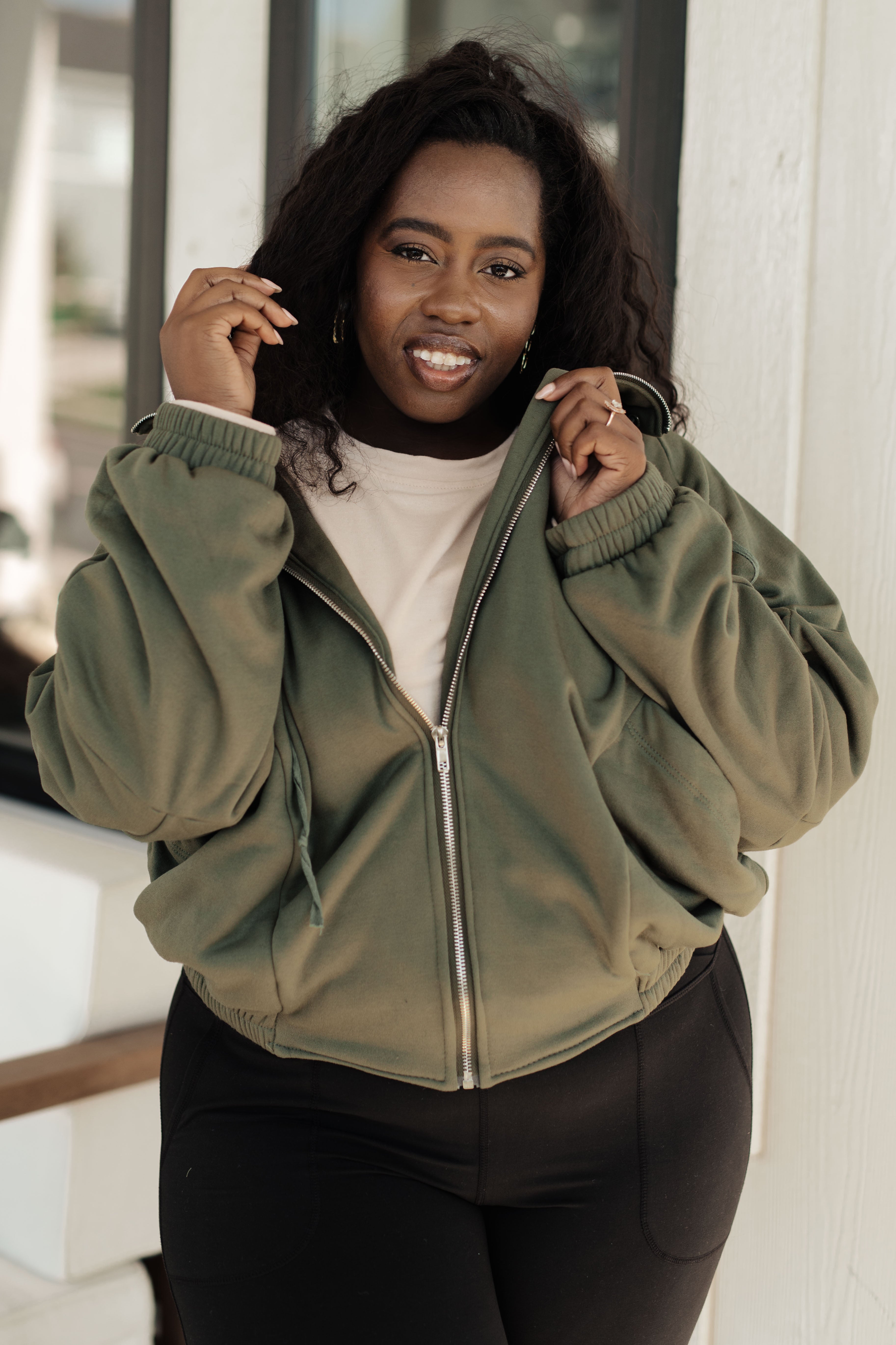 Woman wearing an olive zip-up sweatshirt with relaxed sleeves and elastic cuffs, paired with black pants, standing indoors near a window.