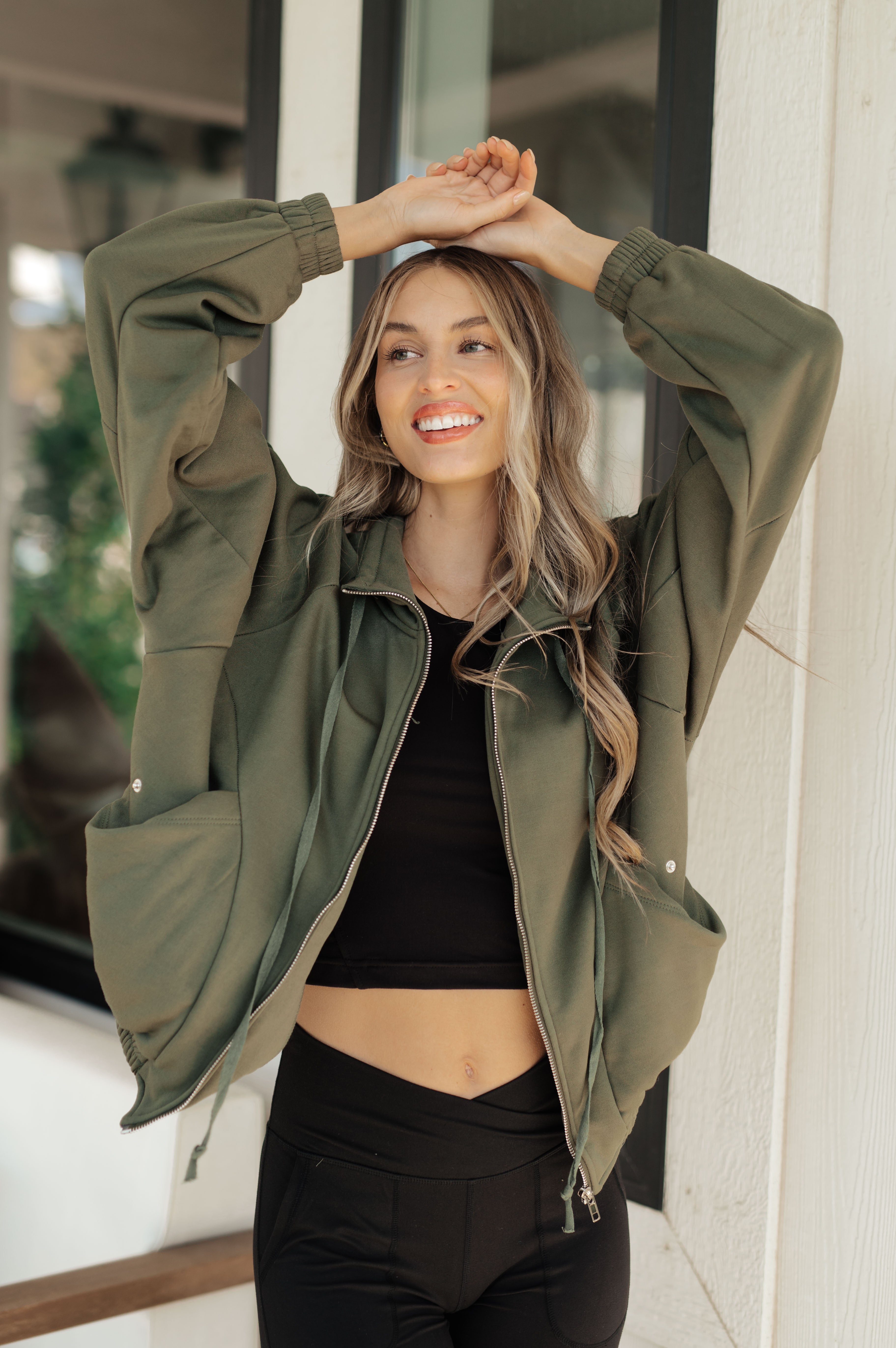 Smiling woman wearing an olive zip-up sweatshirt with gathered cuffs and oversized pockets, paired with black crop top and leggings, standing indoors by a window.