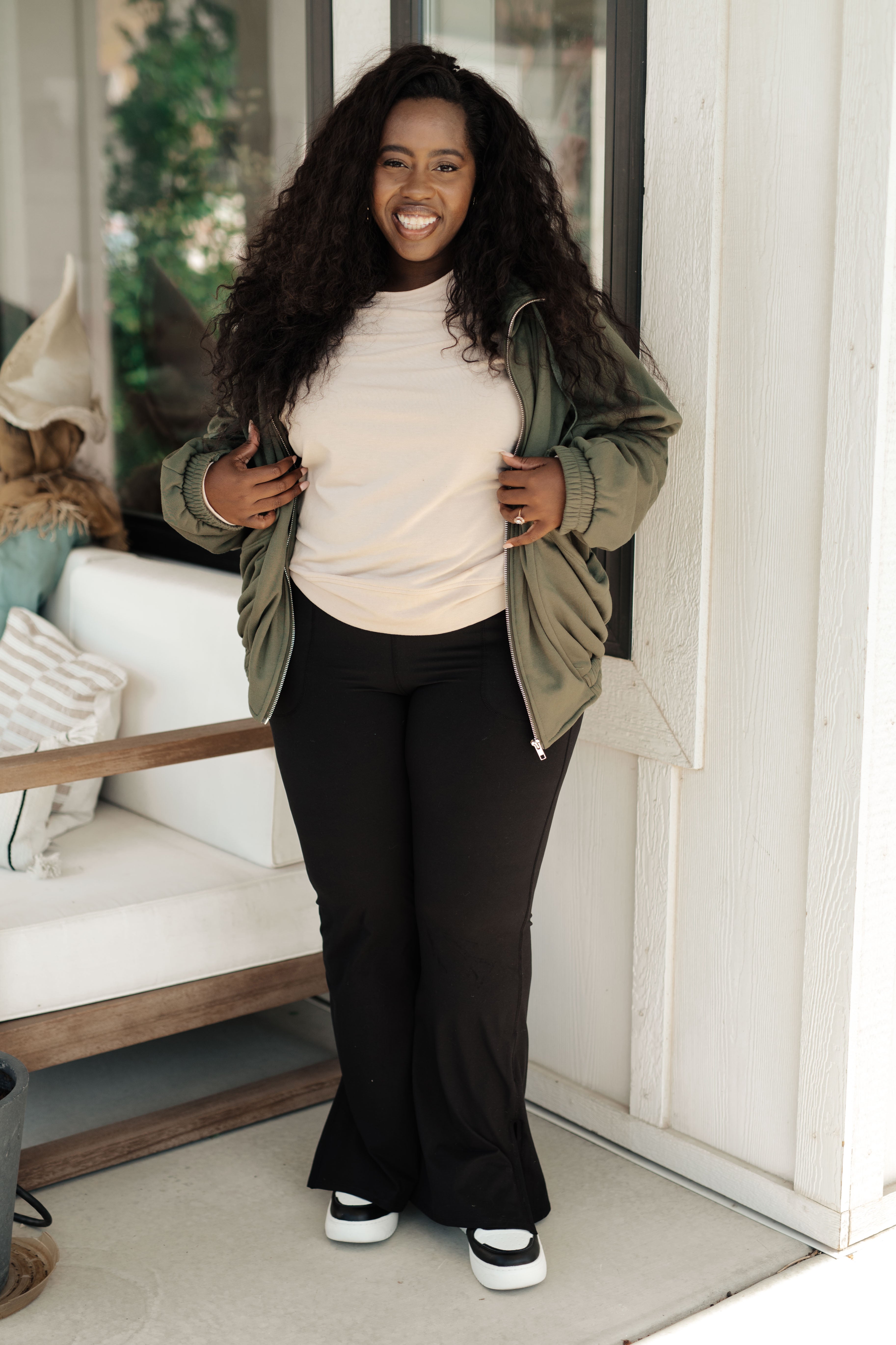 Smiling woman wearing an olive zip-up sweatshirt with elastic cuffs over a light beige top, paired with black flare pants and white sneakers, standing indoors by a window.