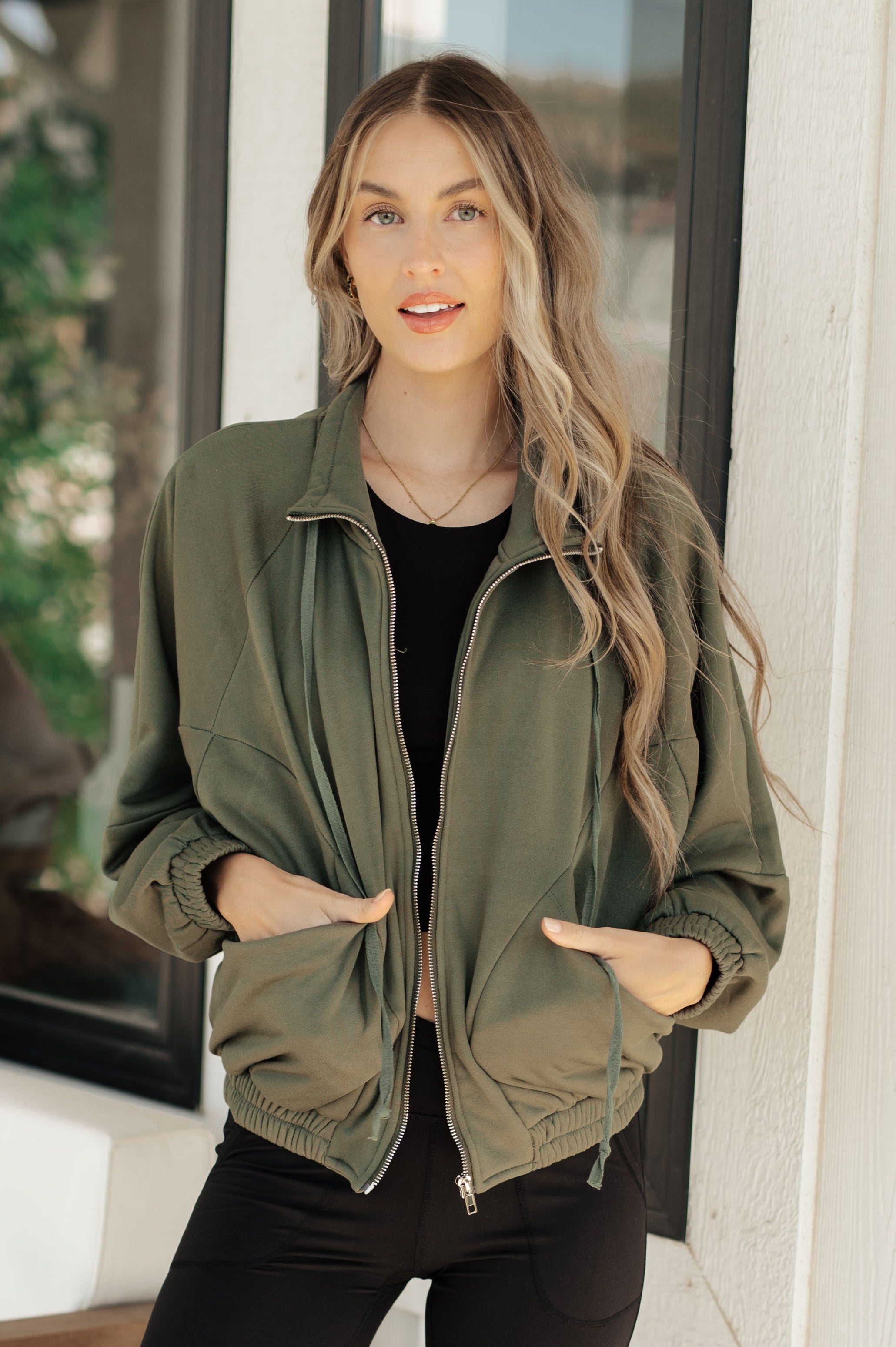 Woman wearing an olive green zip-up sweatshirt with elastic cuffs and hem, paired with black leggings, standing indoors near a window.