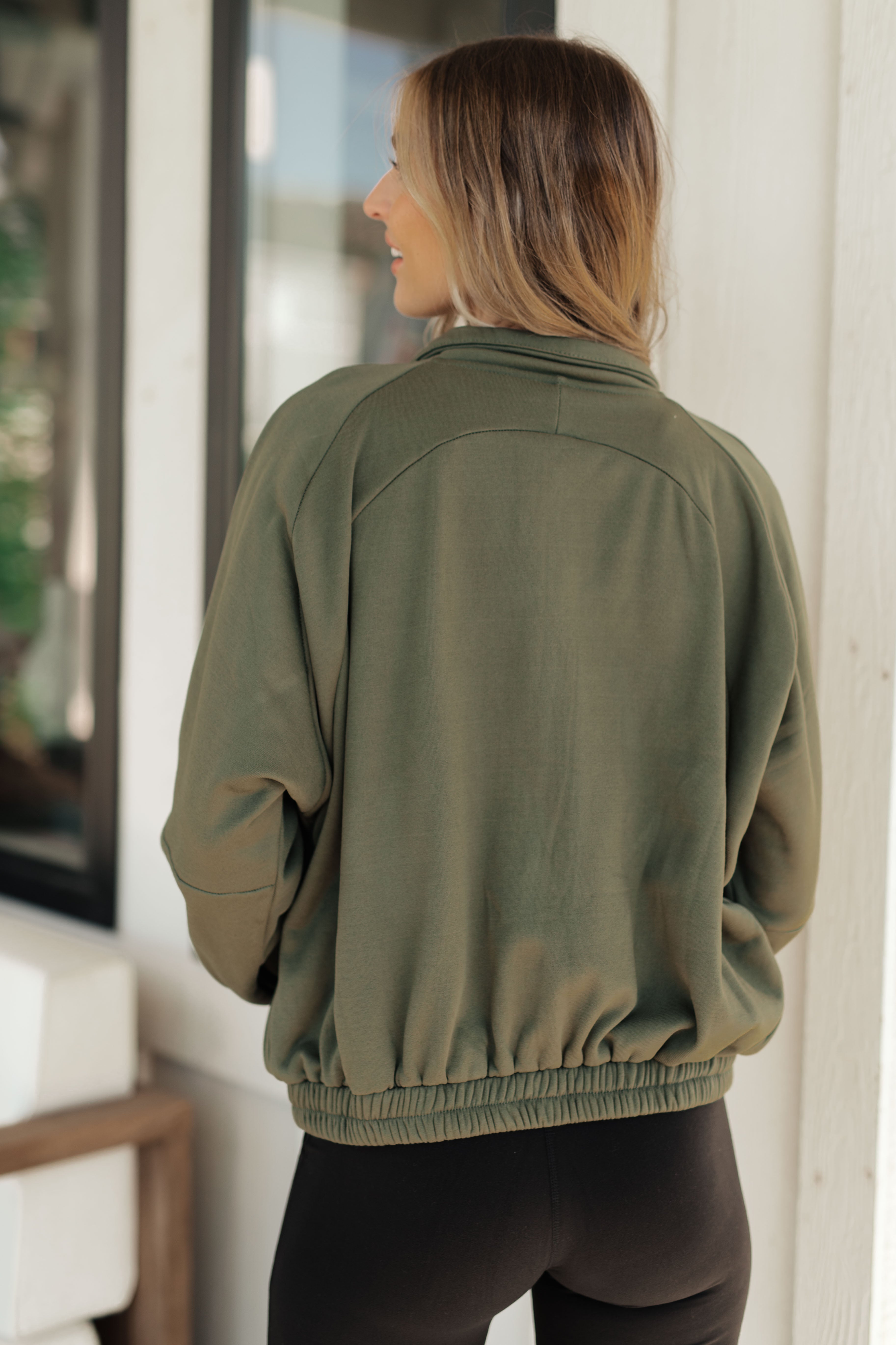 Woman seen from behind wearing an olive zip-up sweatshirt with a relaxed fit and elastic hem, paired with black leggings, standing outdoors near a white wall.