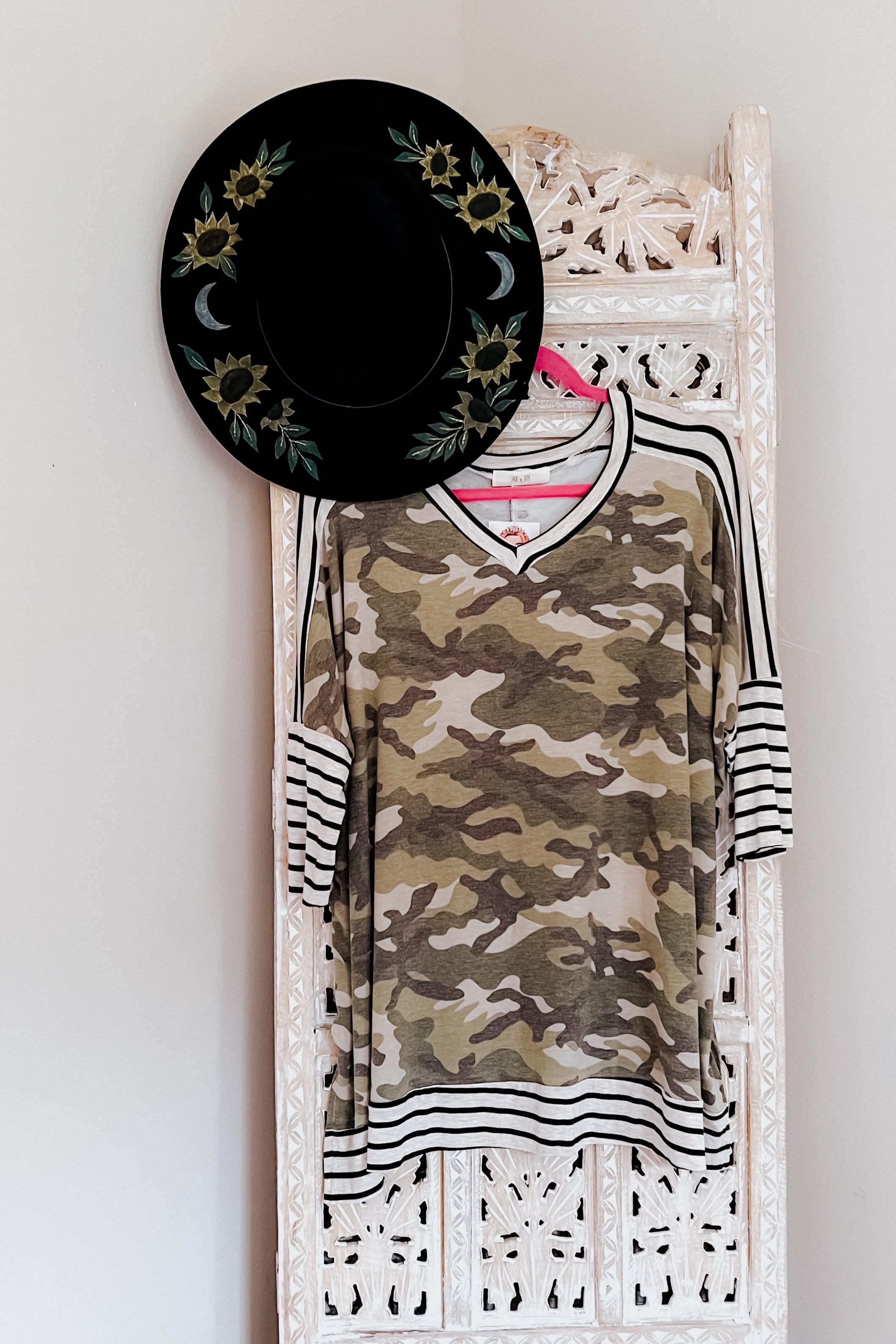 Green camo patterned mid sleeve tunic with contrasting black and white striped sleeves and hem hanging on a pink hanger against a carved white wooden screen, paired with a black hat decorated with sunflowers and crescent moons