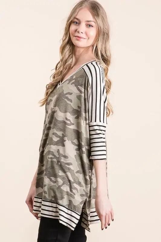 Woman wearing a green camo patterned tunic with black and white striped sleeves and hem, standing against a light background