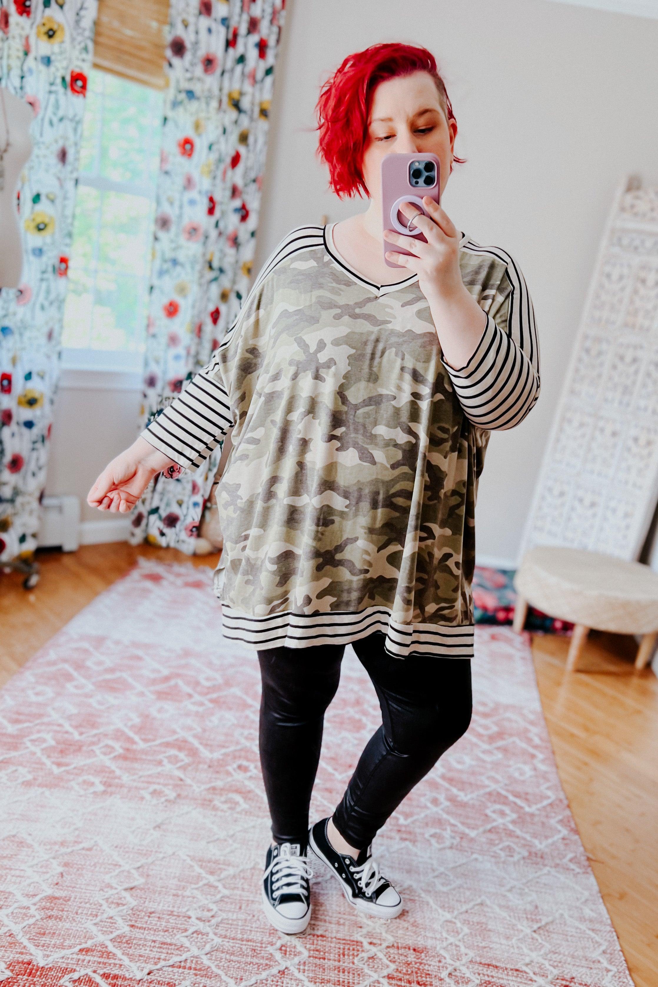 Plus-size woman with bright red hair wearing a green camo patterned tunic featuring striped three-quarter sleeves, neckline, and hem, paired with black leggings and black sneakers, taking a mirror selfie in a cozy room with floral curtains and a patterned rug.