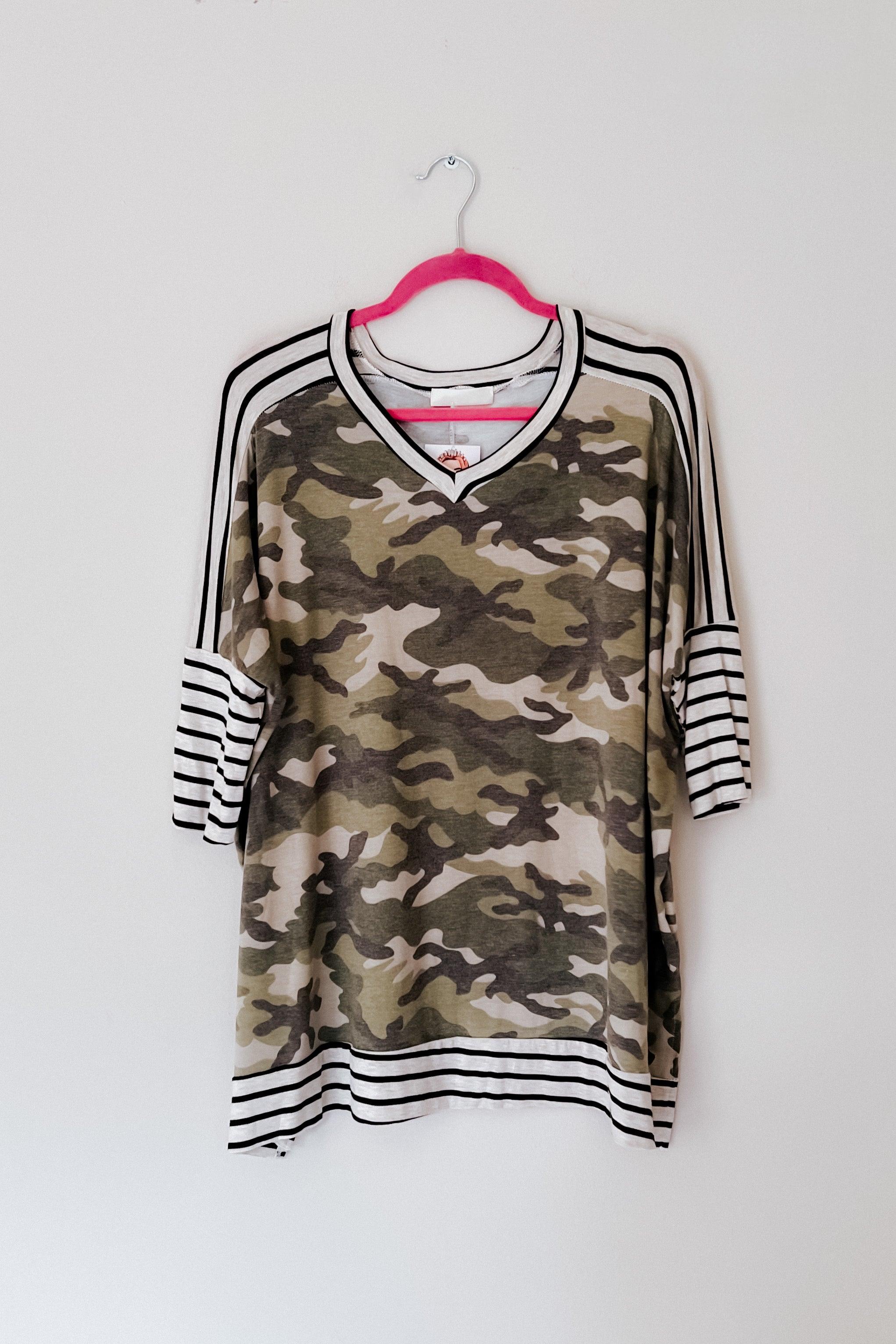 Green camo patterned mid sleeve tunic with contrasting black and white striped sleeves, cuffs, neckline, and hem hanging on a pink hanger against a white background