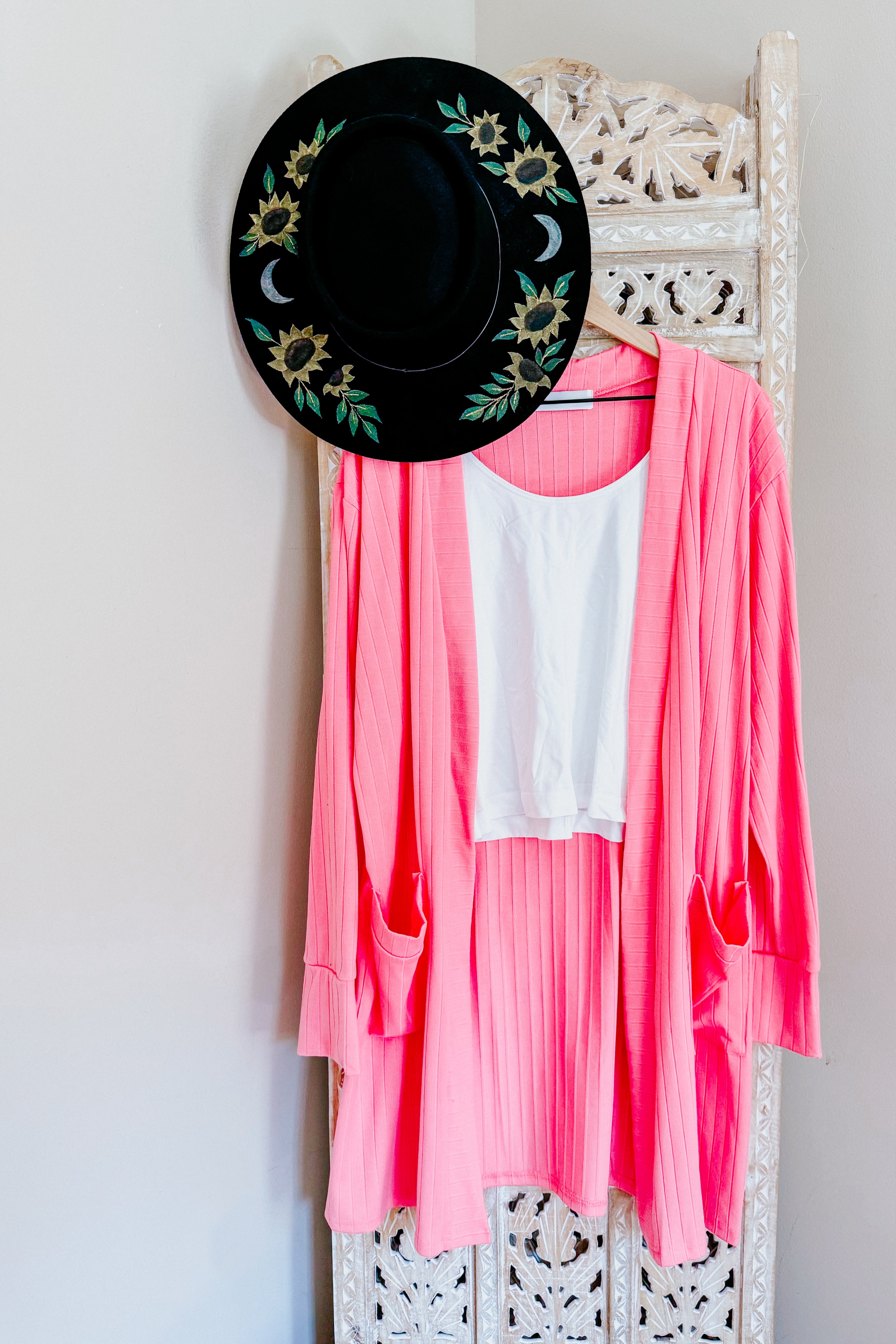 Bright pink Modern Idealist open front cardigan hanging with white tank top and black embroidered hat on carved screen