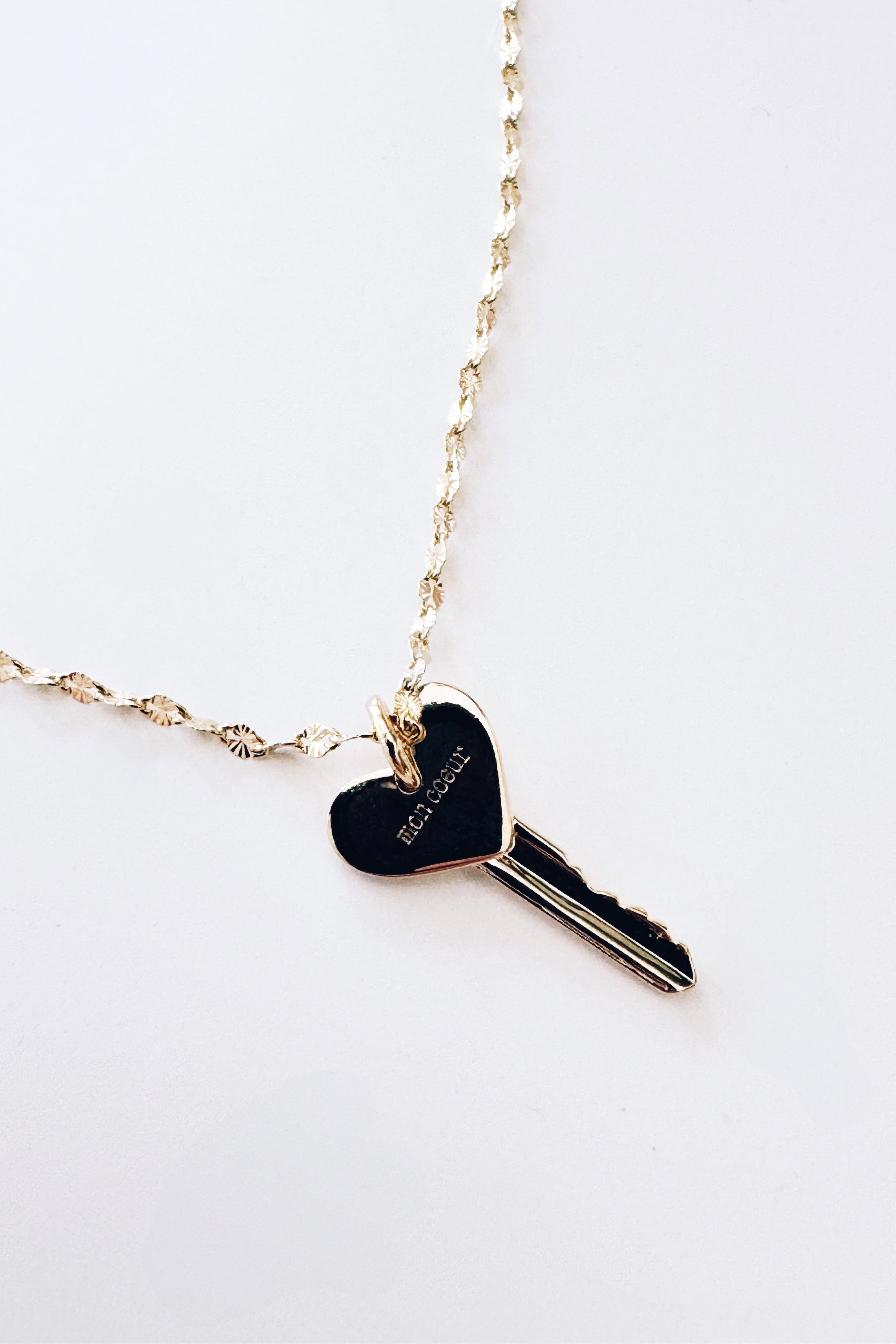 Gold necklace featuring a heart-shaped key pendant with engraved lettering on a delicate textured chain against a white background.