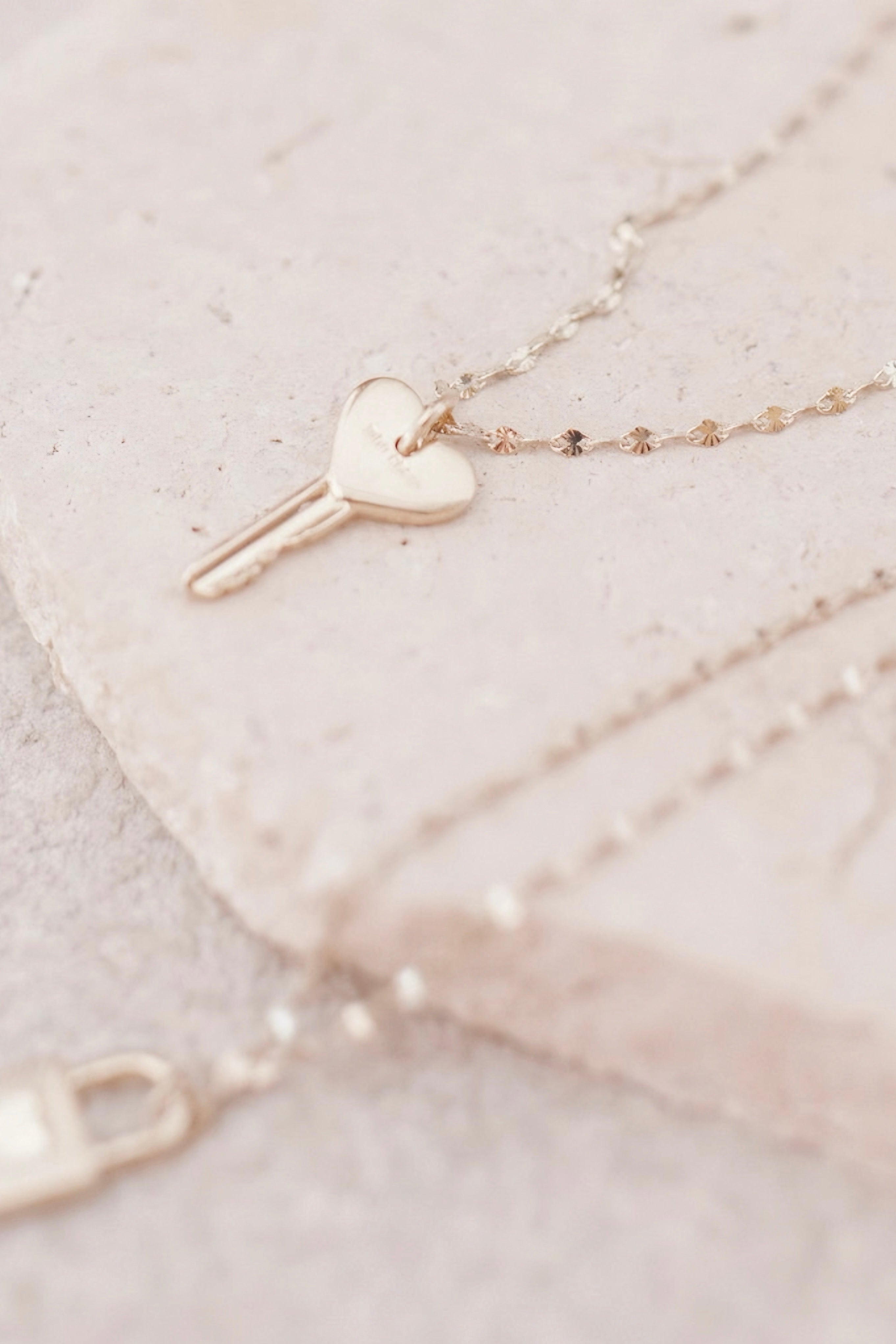 Delicate gold necklace with a heart-shaped key pendant resting on a textured light stone surface, highlighting the fine chain details.