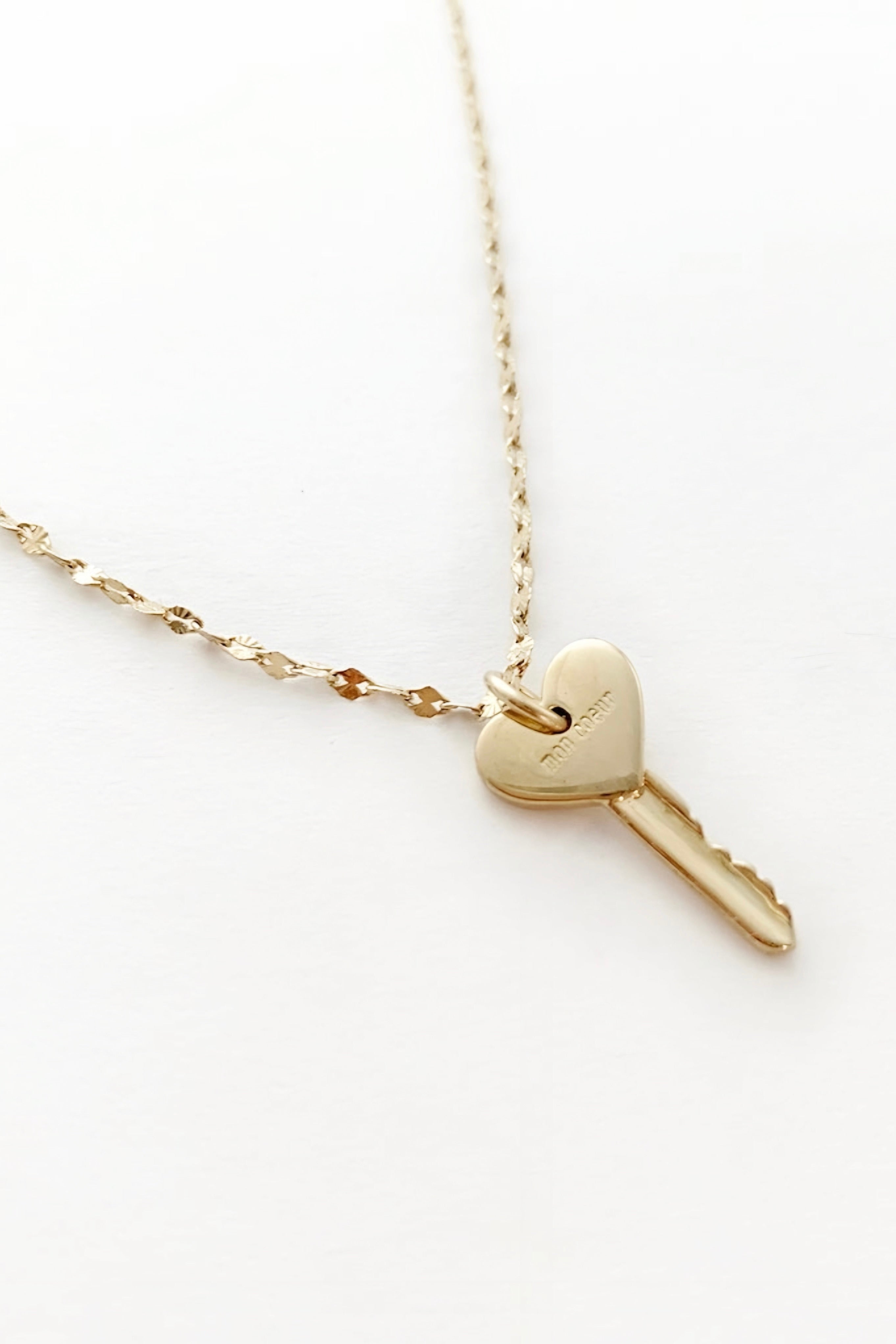 Elegant gold necklace featuring a heart-shaped key pendant with delicate engraving on a fine textured chain against a white background.