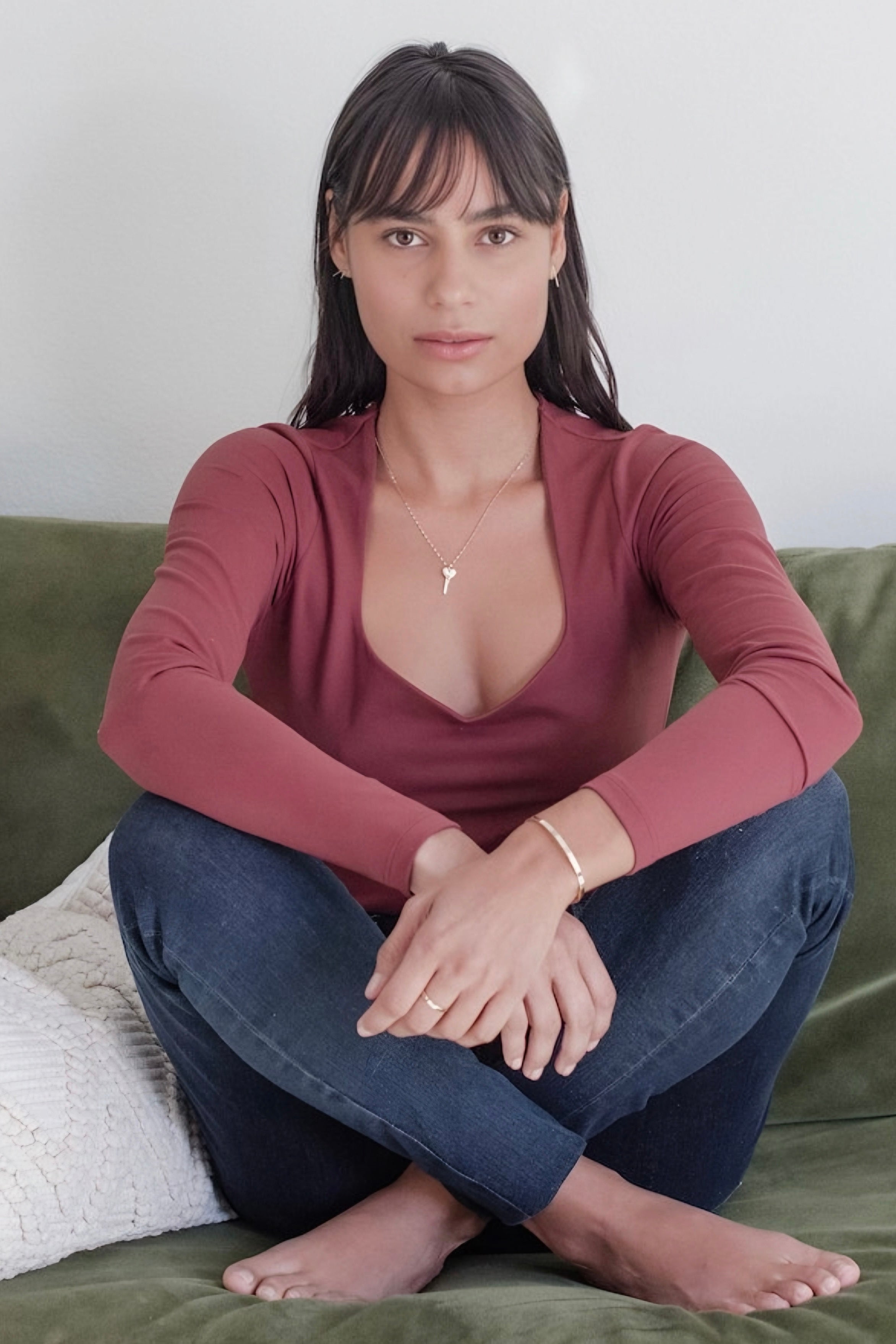 Woman seated cross-legged on a green sofa wearing a deep mauve long-sleeve top, dark jeans, a gold key pendant necklace, and minimal jewelry.