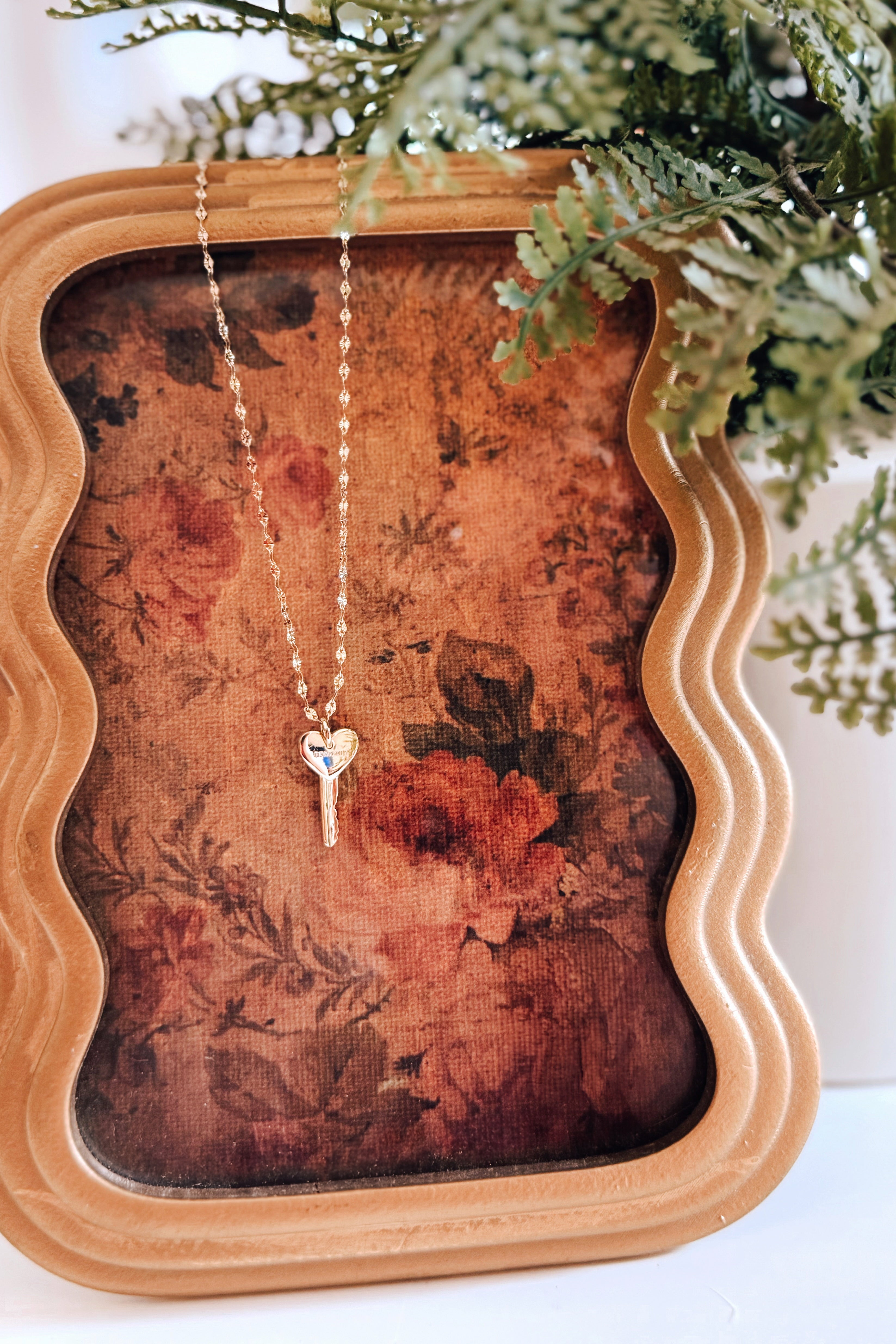 Gold heart-shaped key pendant necklace displayed on a vintage floral fabric background inside a decorative wooden frame with greenery nearby.