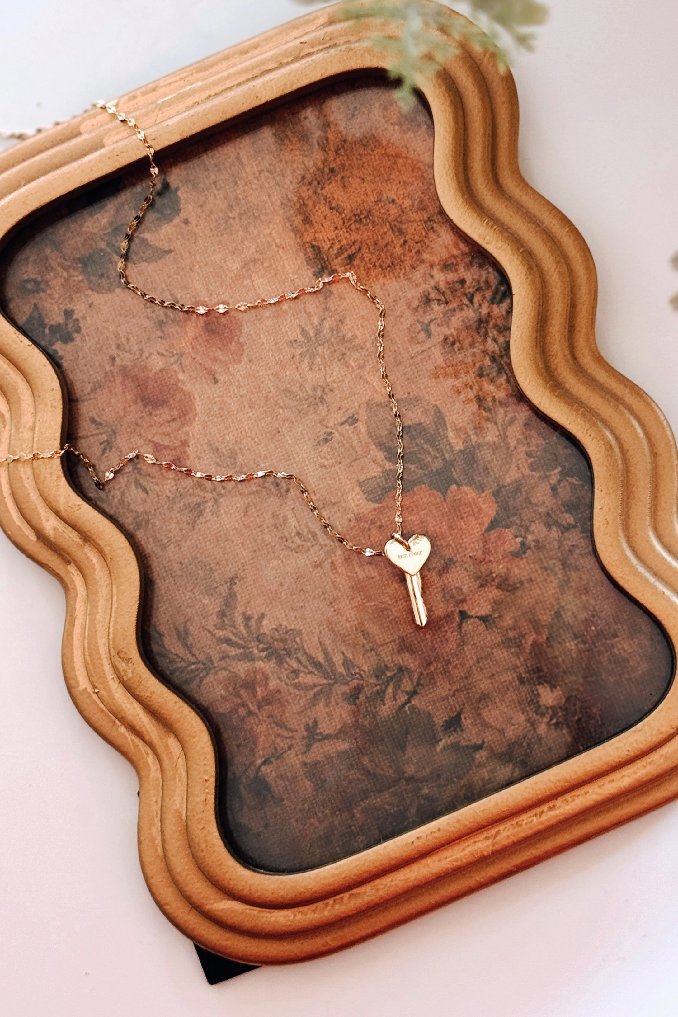 Gold key necklace with a heart-shaped pendant resting on a vintage floral-patterned fabric inside an ornate wooden frame.