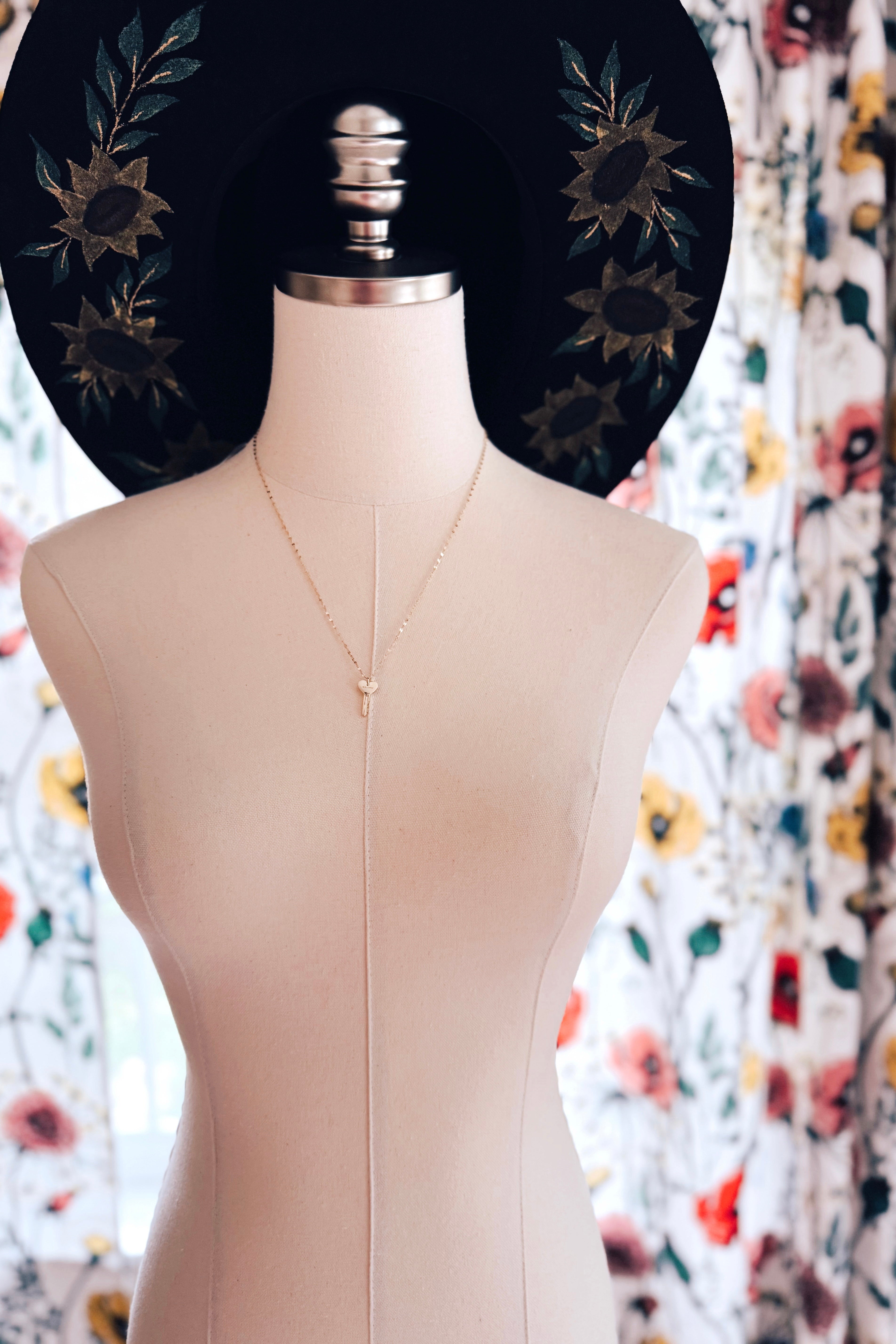 Gold key necklace with a small heart-shaped pendant displayed on a mannequin bust against a floral curtain background and a black hat with floral embroidery.