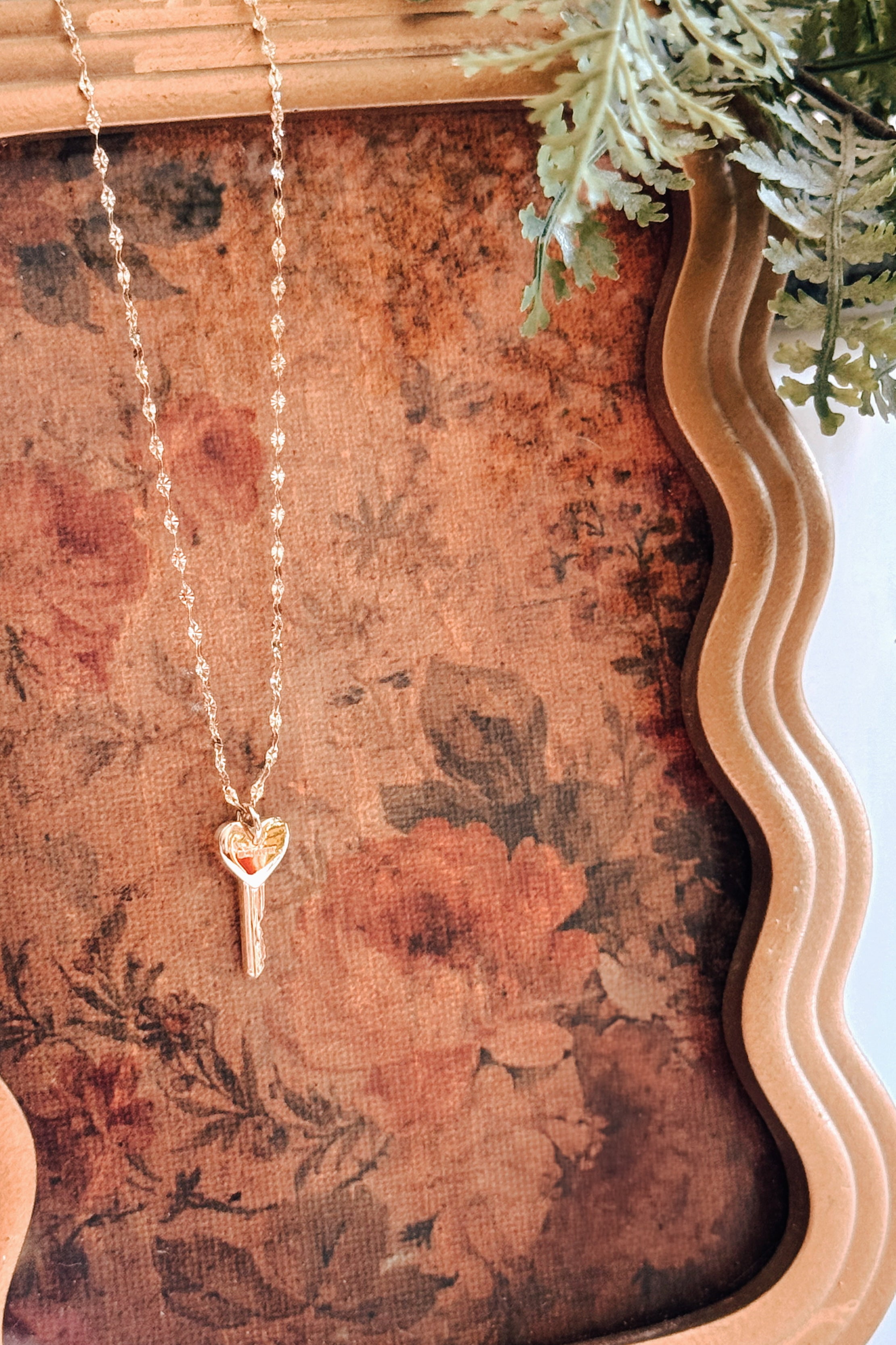 Gold key pendant with a heart-shaped top hanging from a delicate chain, displayed on a floral-patterned vintage fabric inside a decorative frame.