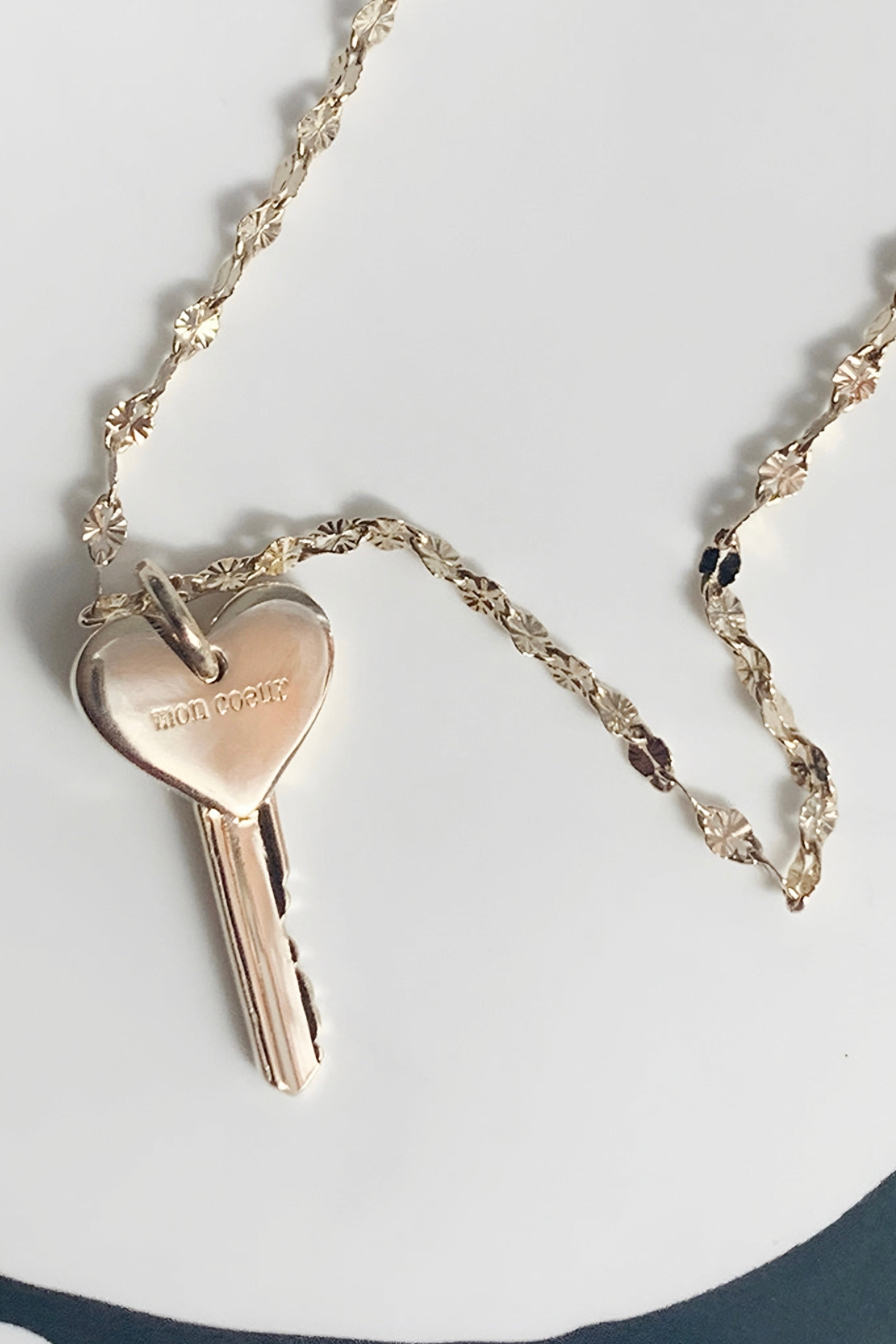 Gold key pendant with a heart-shaped top engraved with "mon coeur," hanging from a textured gold chain on a white background.
