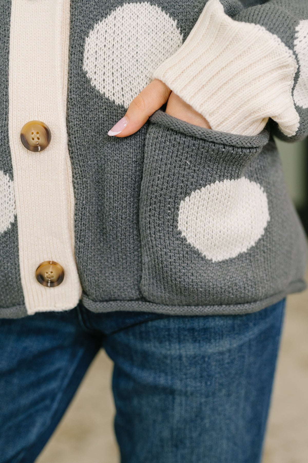 Close-up of a charcoal knit cardigan with oversized cream polka dots and a front patch pocket paired with blue jeans