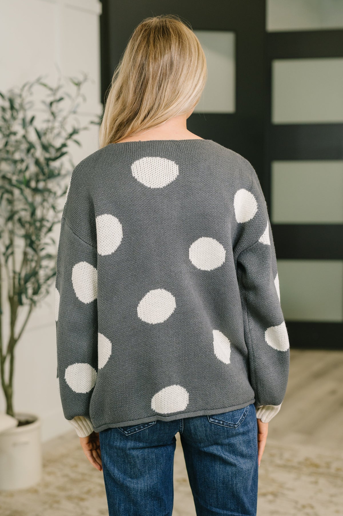 Woman seen from behind wearing a charcoal knit cardigan with oversized cream polka dots and blue jeans indoors