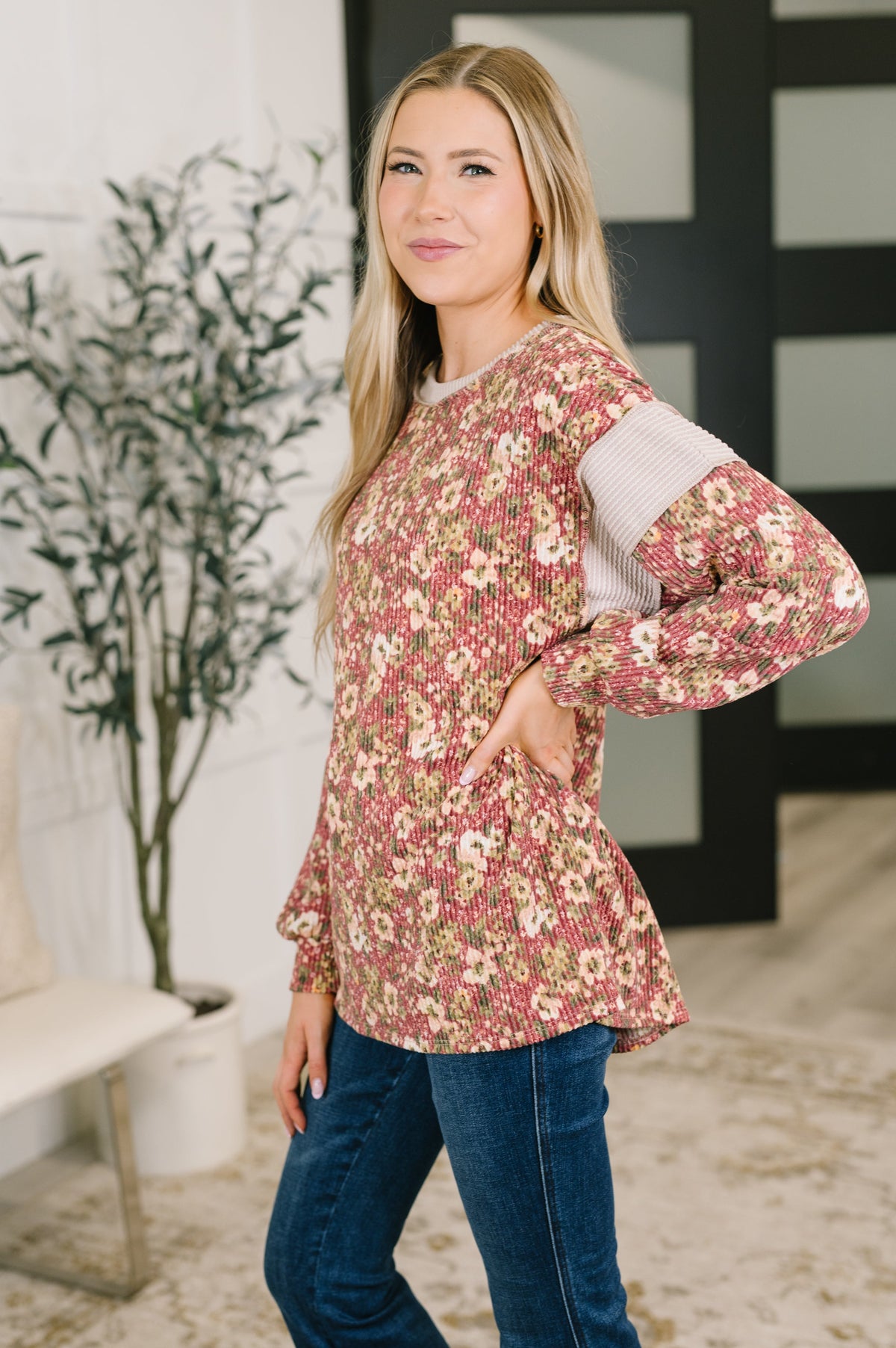 Woman wearing a rust floral textured long sleeve top with contrasting shoulder panels and dark jeans indoors