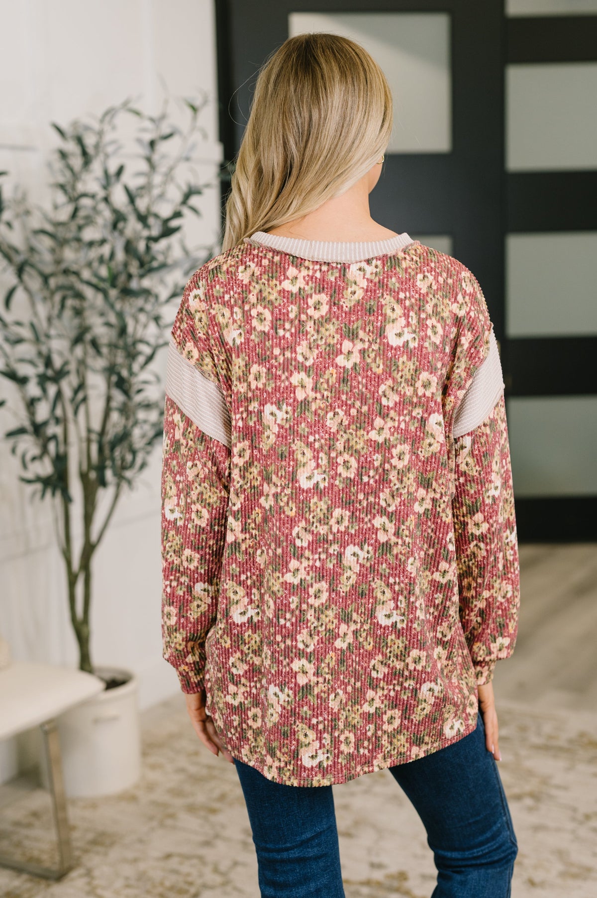 Back view of a woman wearing a rust floral textured knit top with contrasting shoulder panels and dark jeans indoors beside a plant