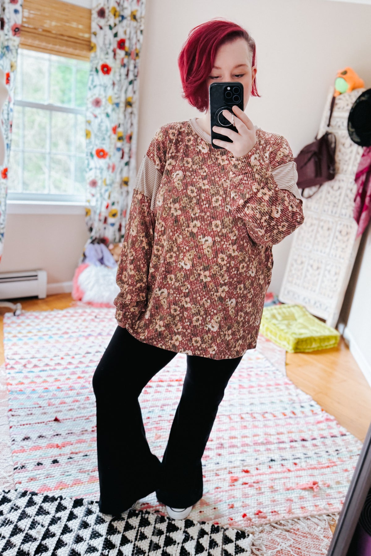 Red haired woman taking a bedroom mirror selfie in a rust textured floral top with relaxed sleeves black pants and white sneakers