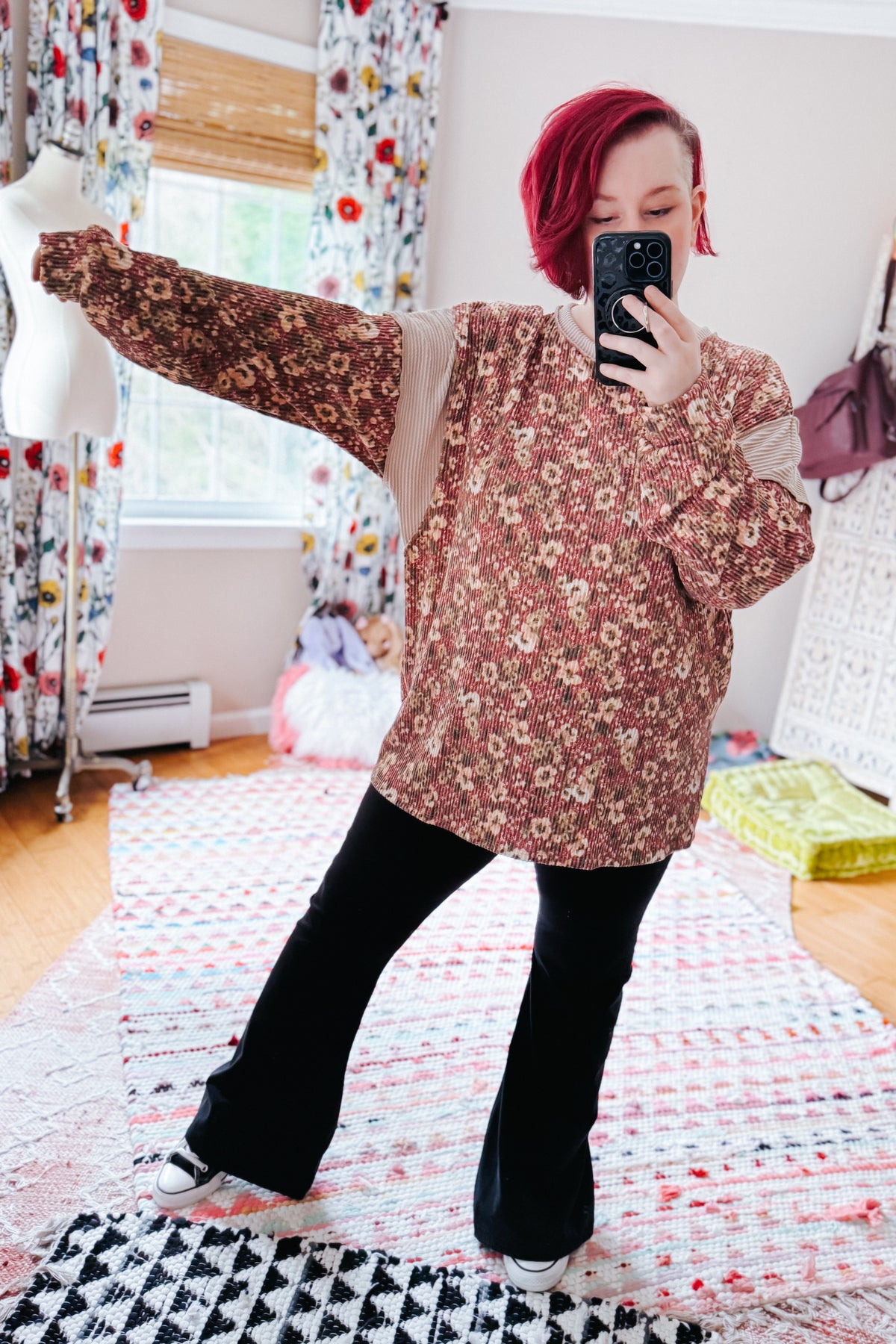 Red haired woman taking a mirror selfie in a rust textured long sleeve top with black flared pants and sneakers in a colorful bedroom