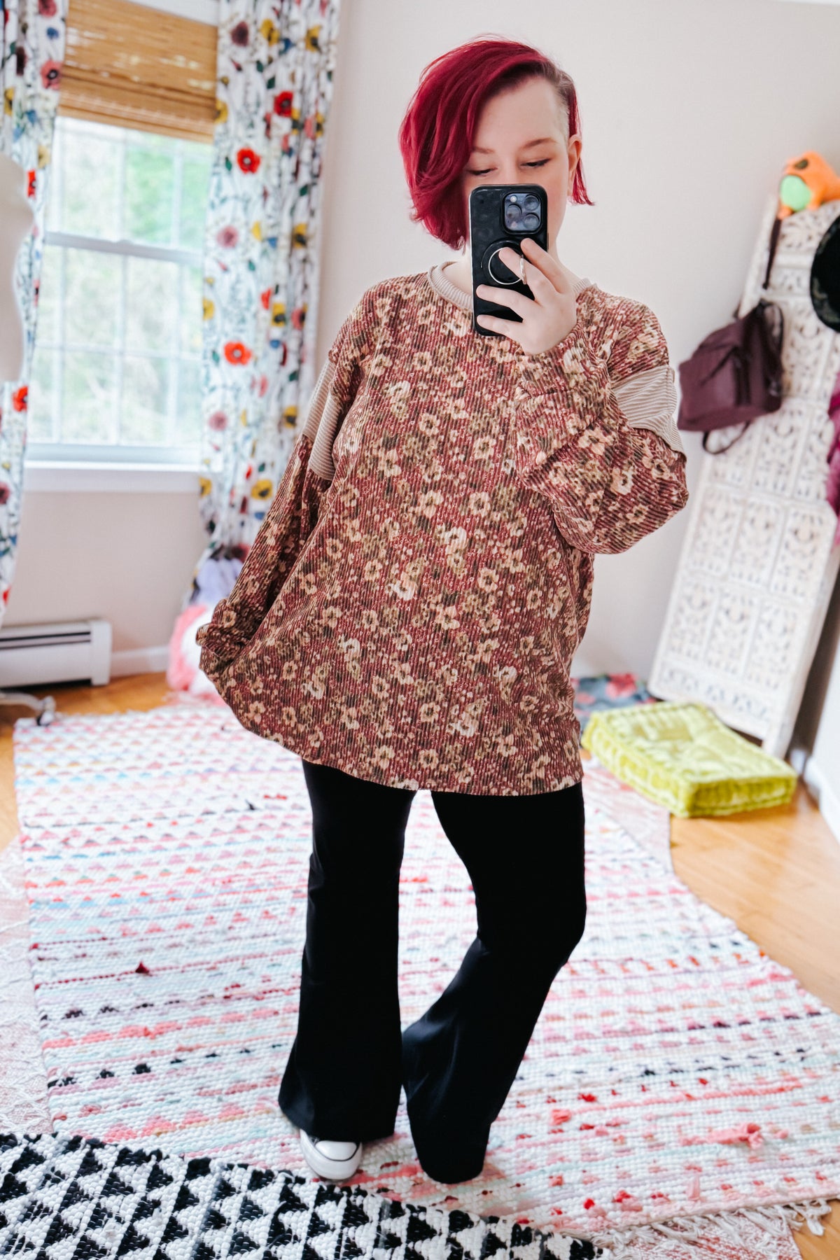 Red haired woman taking a bedroom mirror selfie in a rust textured long sleeve top with black pants and white sneakers