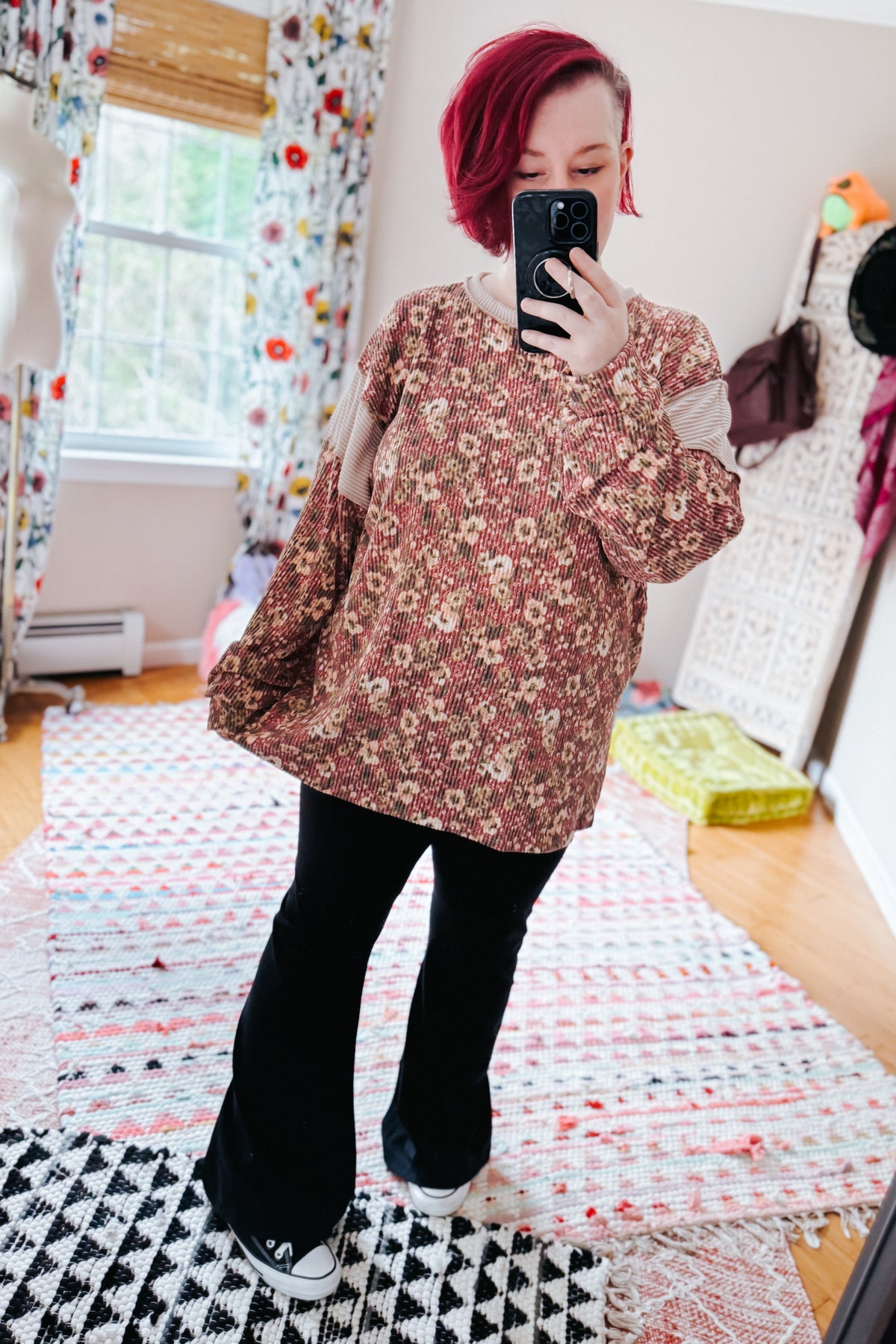 Red haired woman taking a bedroom mirror selfie in a rust textured floral long sleeve top with black flared pants and sneakers