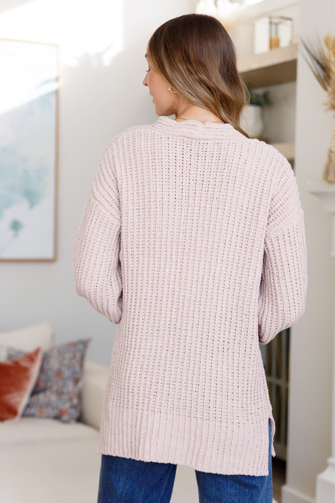 Woman wearing a light pink button down cardigan and blue jeans standing in an indoor setting.