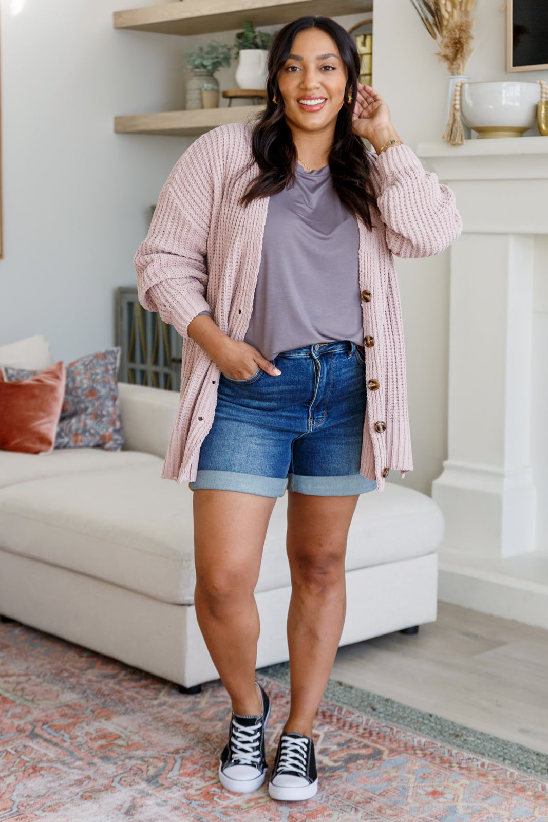 Woman wearing a light pink button down cardigan over a grey top, blue jean shorts and black converse sneakers standing in an indoor setting.