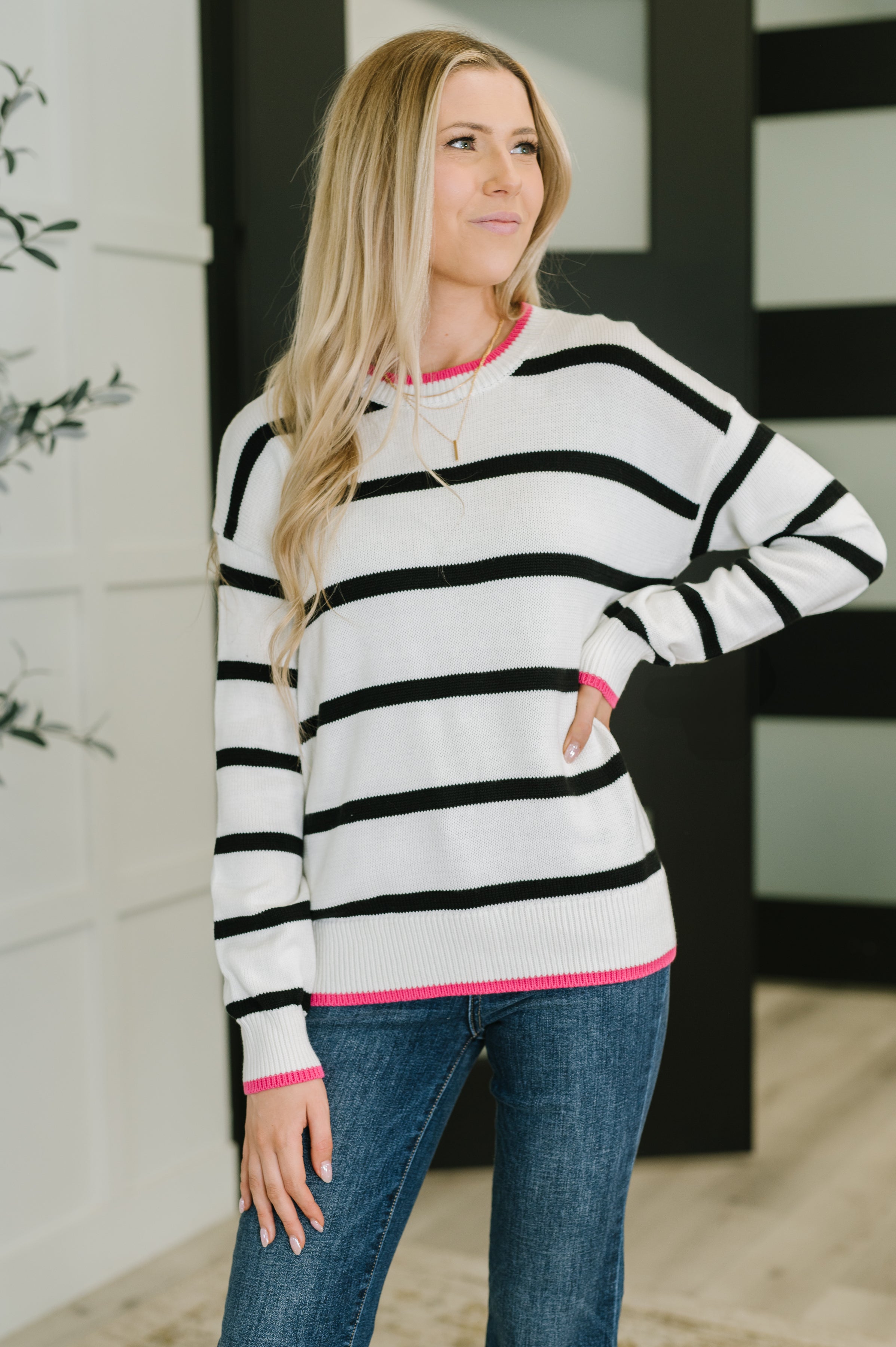 Woman wearing an ivory lightweight sweater with black horizontal stripes and bright pink trim at the collar, cuffs, and hem, paired with blue jeans.