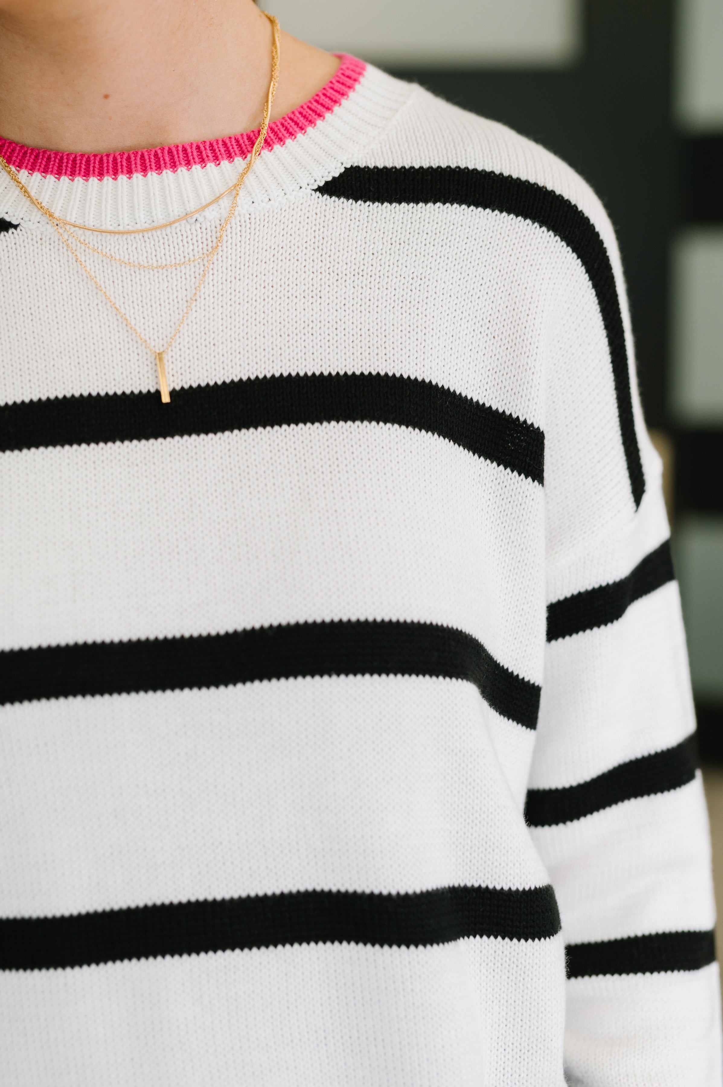 Close-up of an ivory sweater with horizontal black stripes and bright pink trim at the neckline, paired with layered delicate gold necklaces.