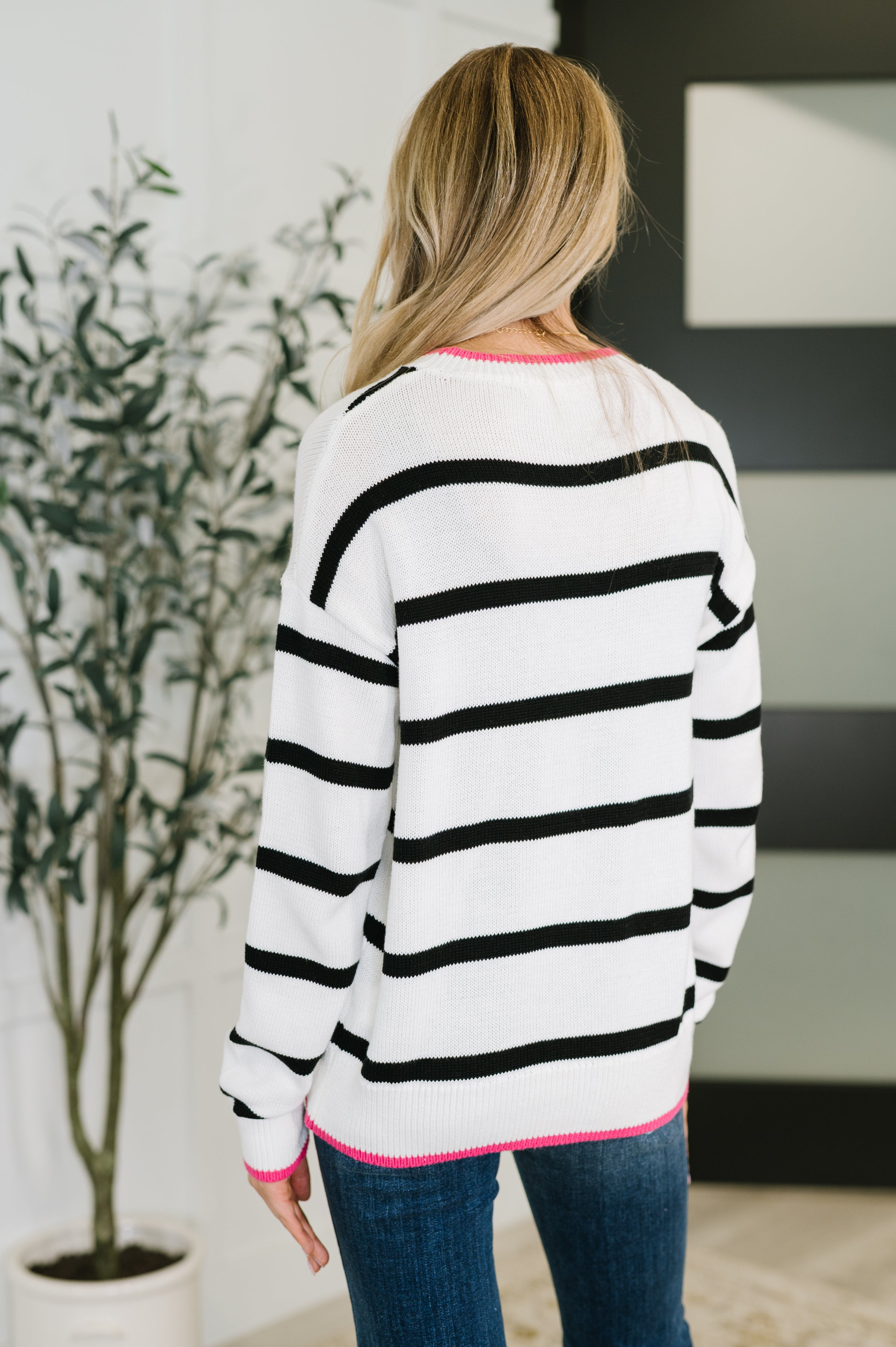 Back view of a woman wearing a lightweight ivory sweater with black stripes and bright pink edging at the collar, cuffs, and hem, paired with blue jeans.