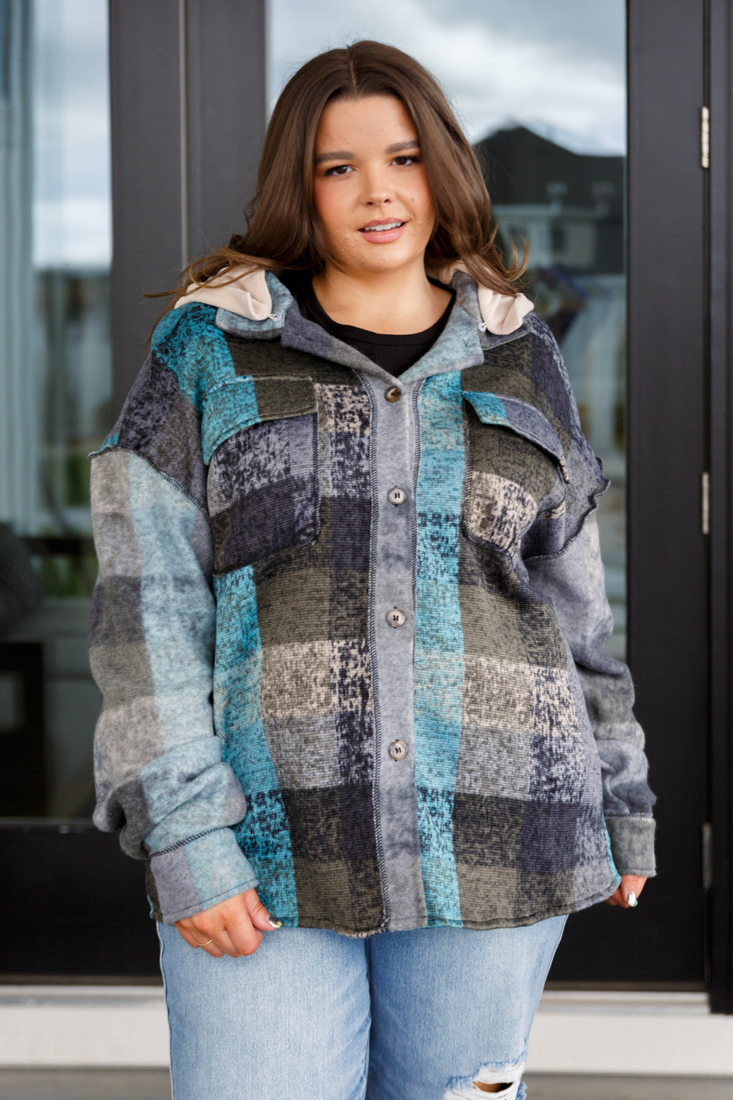 Woman wearing a blue and gray plaid hooded shacket with button front and chest pockets, paired with light blue distressed jeans.