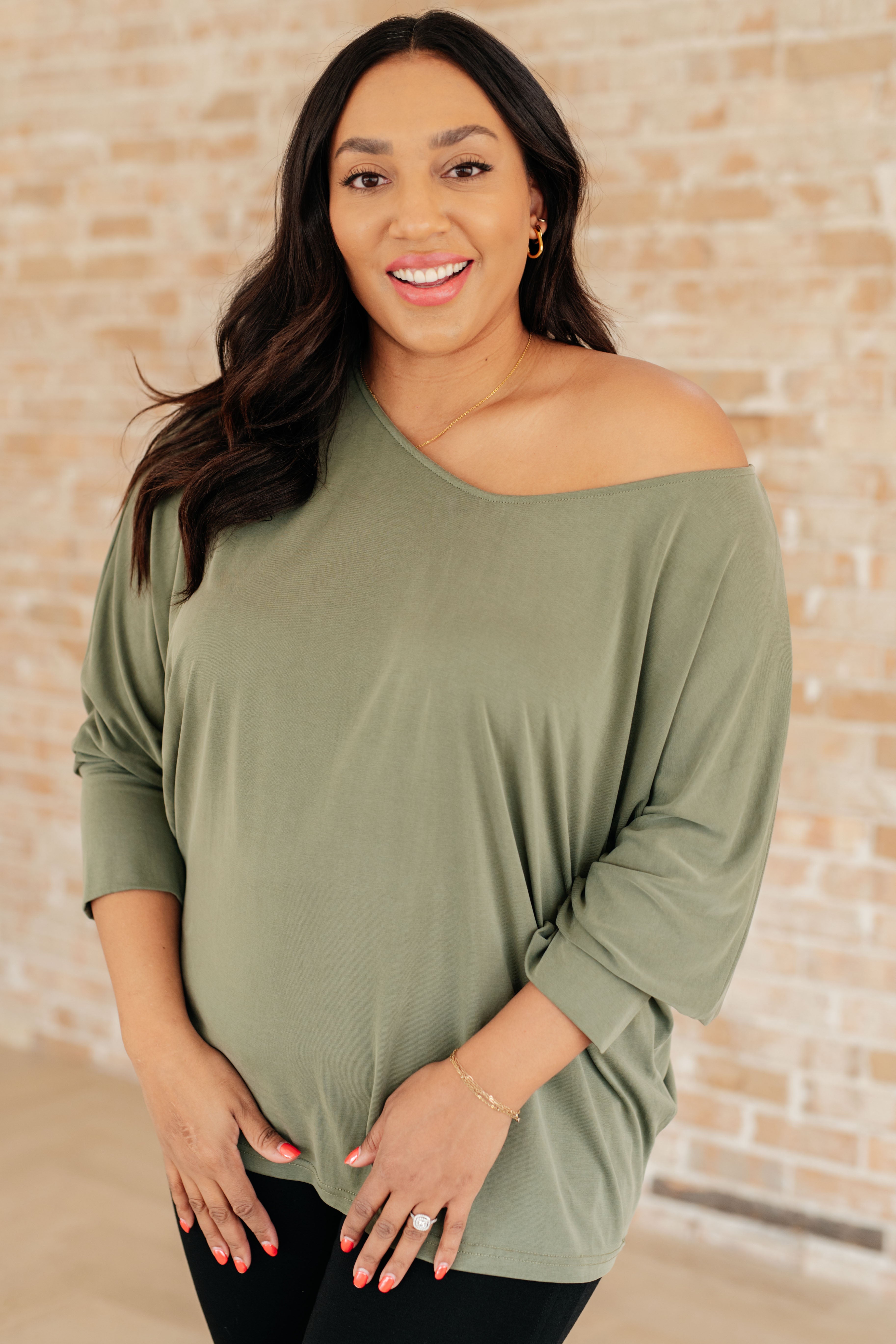 Woman modeling a green long sleeve top with a boat neckline and relaxed dolman sleeves over black pants indoors