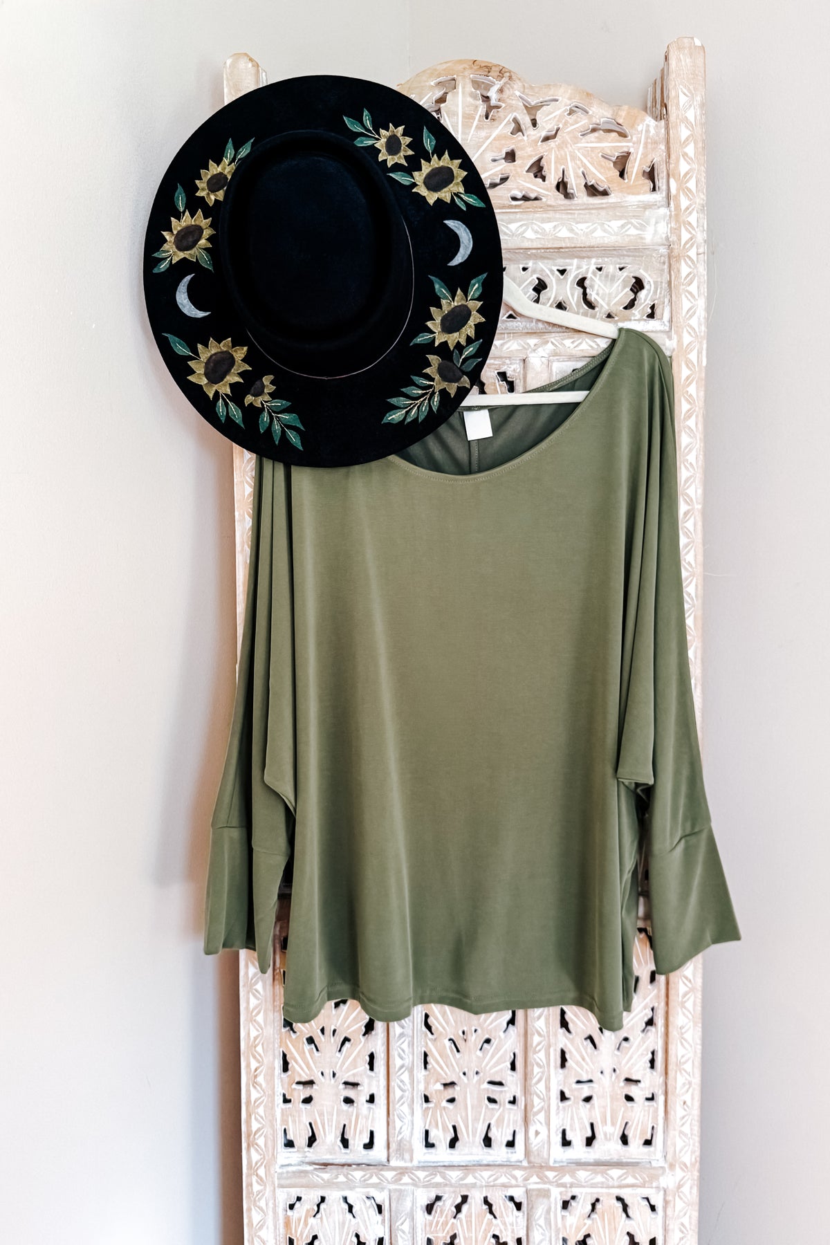 Green long sleeve top with a boat neckline dolman sleeves and flared cuffs hanging beneath a black floral hat on an ornate white rack