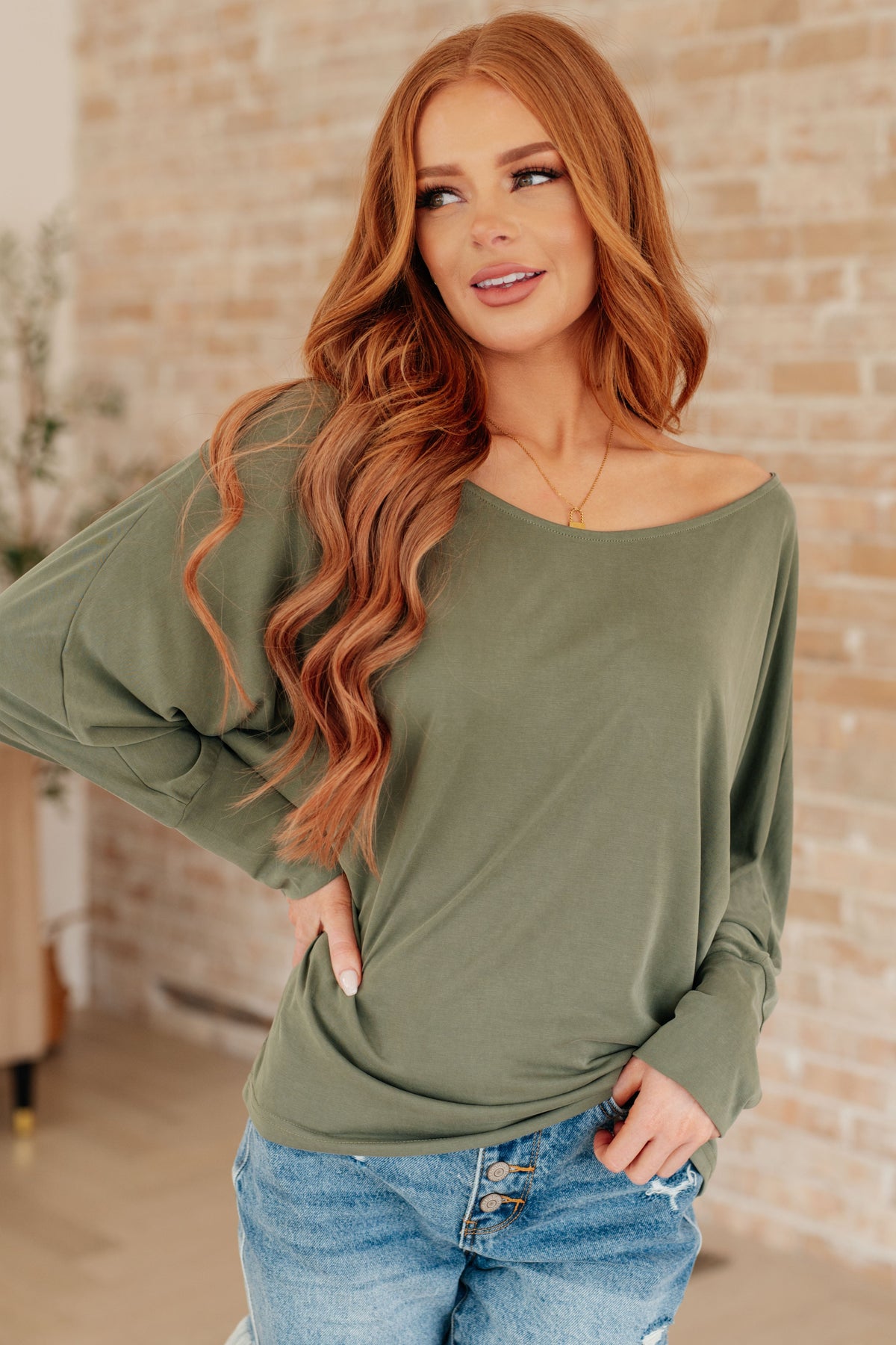 Woman modeling a green long sleeve top with a boat neckline and dolman sleeves over distressed blue jeans in a brick walled room