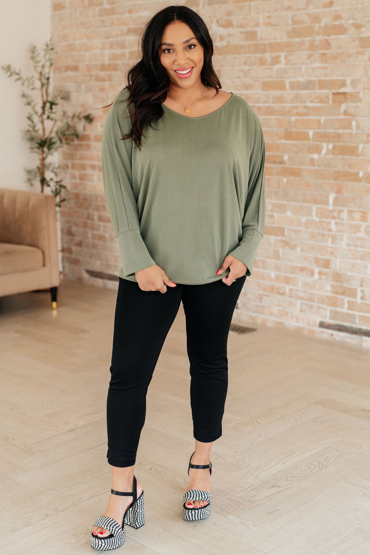 Woman modeling a green long sleeve top with dolman sleeves and extended cuffs black pants and striped heels indoors