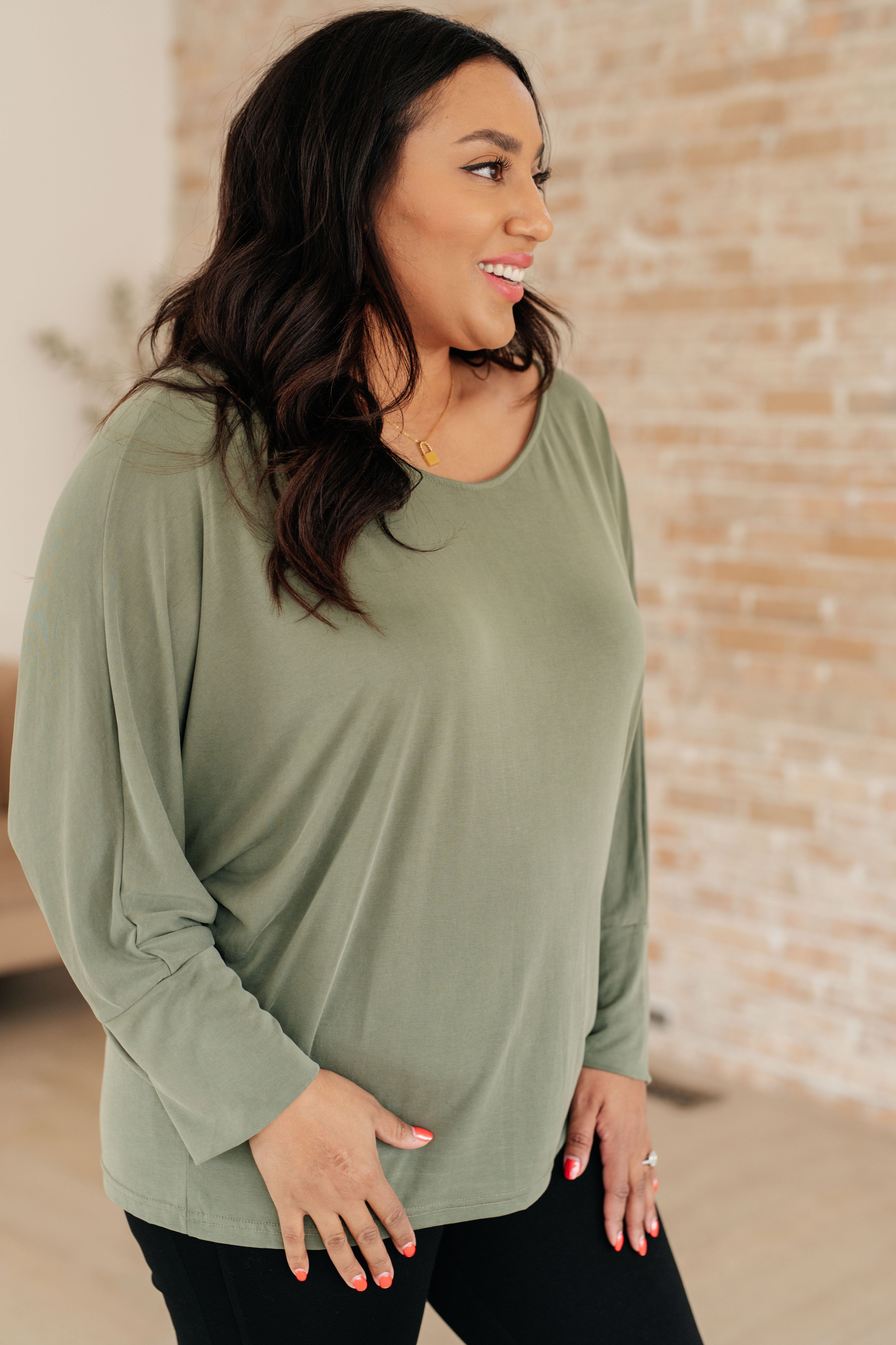Smiling woman wearing a green boat neck long sleeve top with relaxed dolman sleeves and black pants in a bright brick walled room