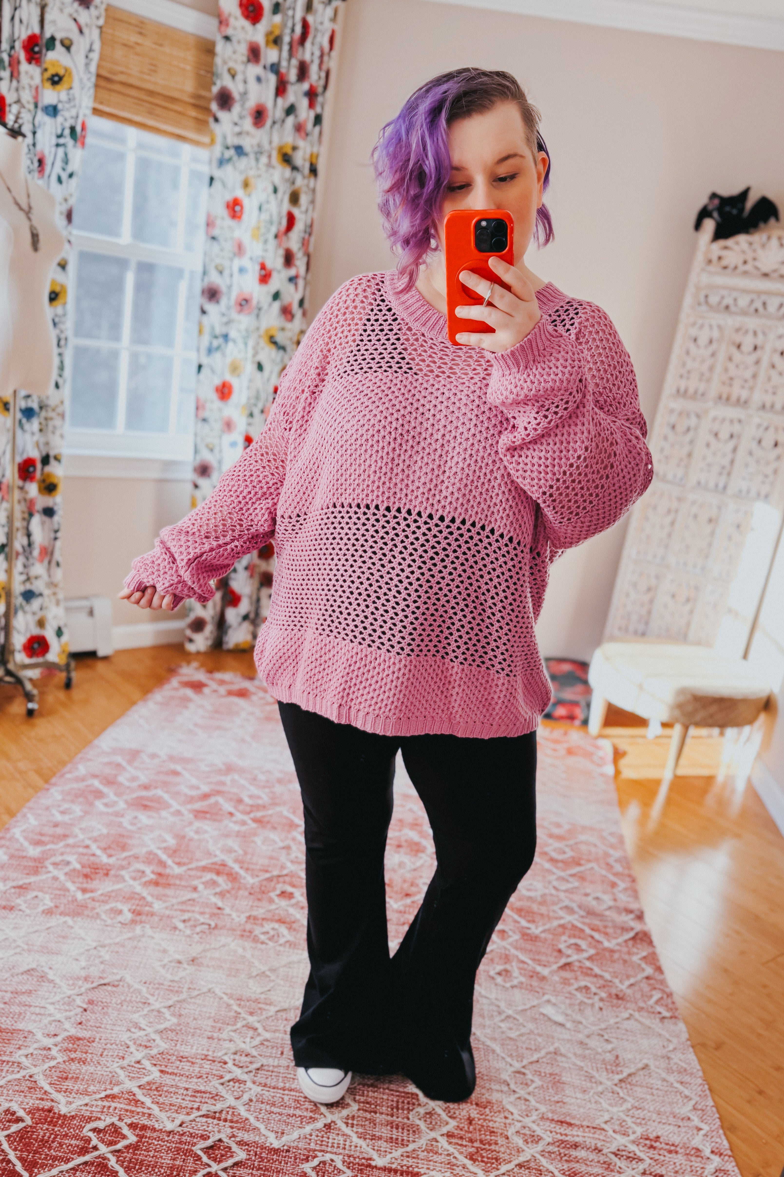Woman wearing a loose pink knit sweater with open weave and black pants taking a mirror selfie indoors
