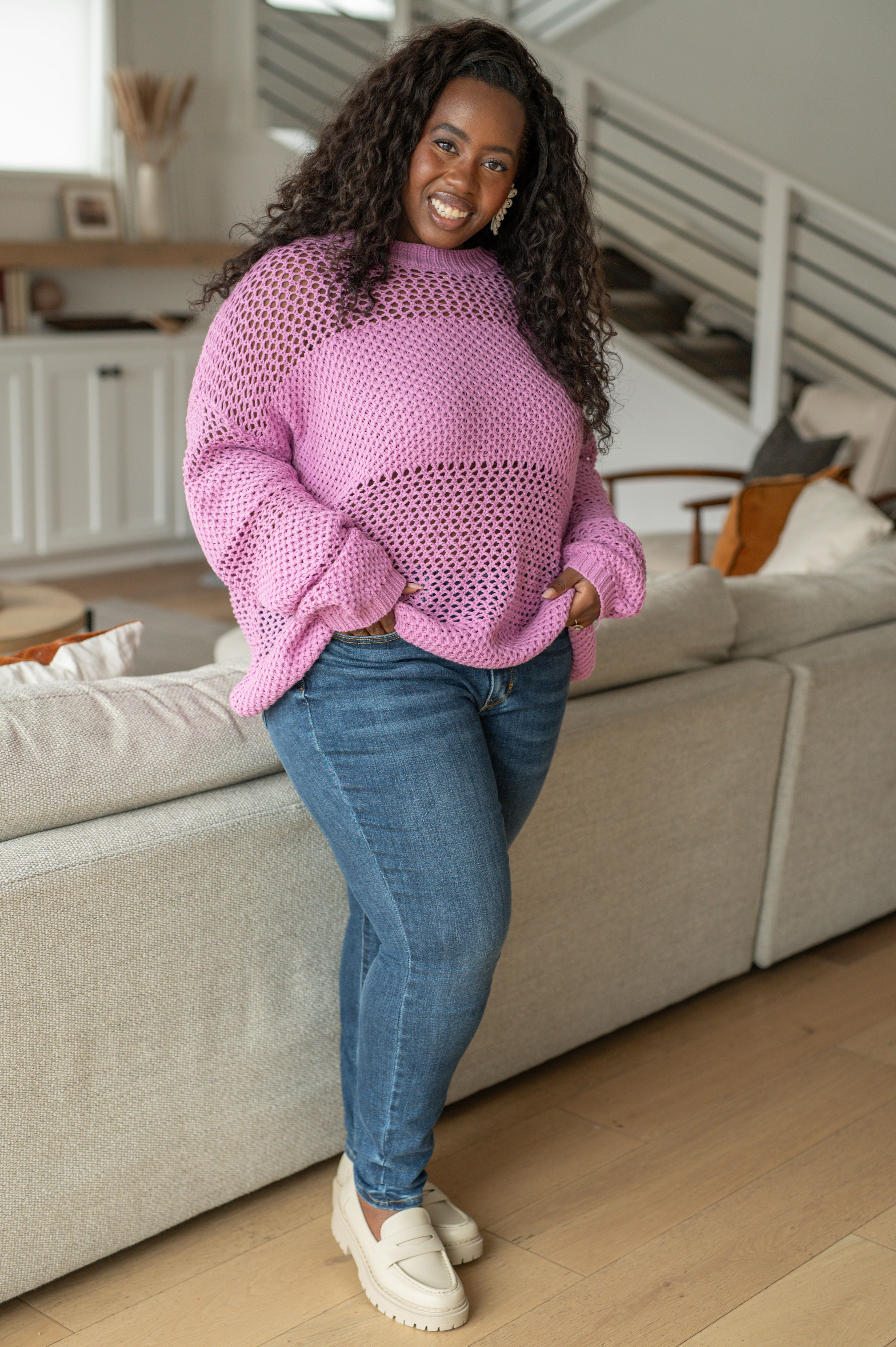Plus-size woman smiling indoors wearing a loose lavender knit sweater with open weave paired with blue jeans and cream loafers