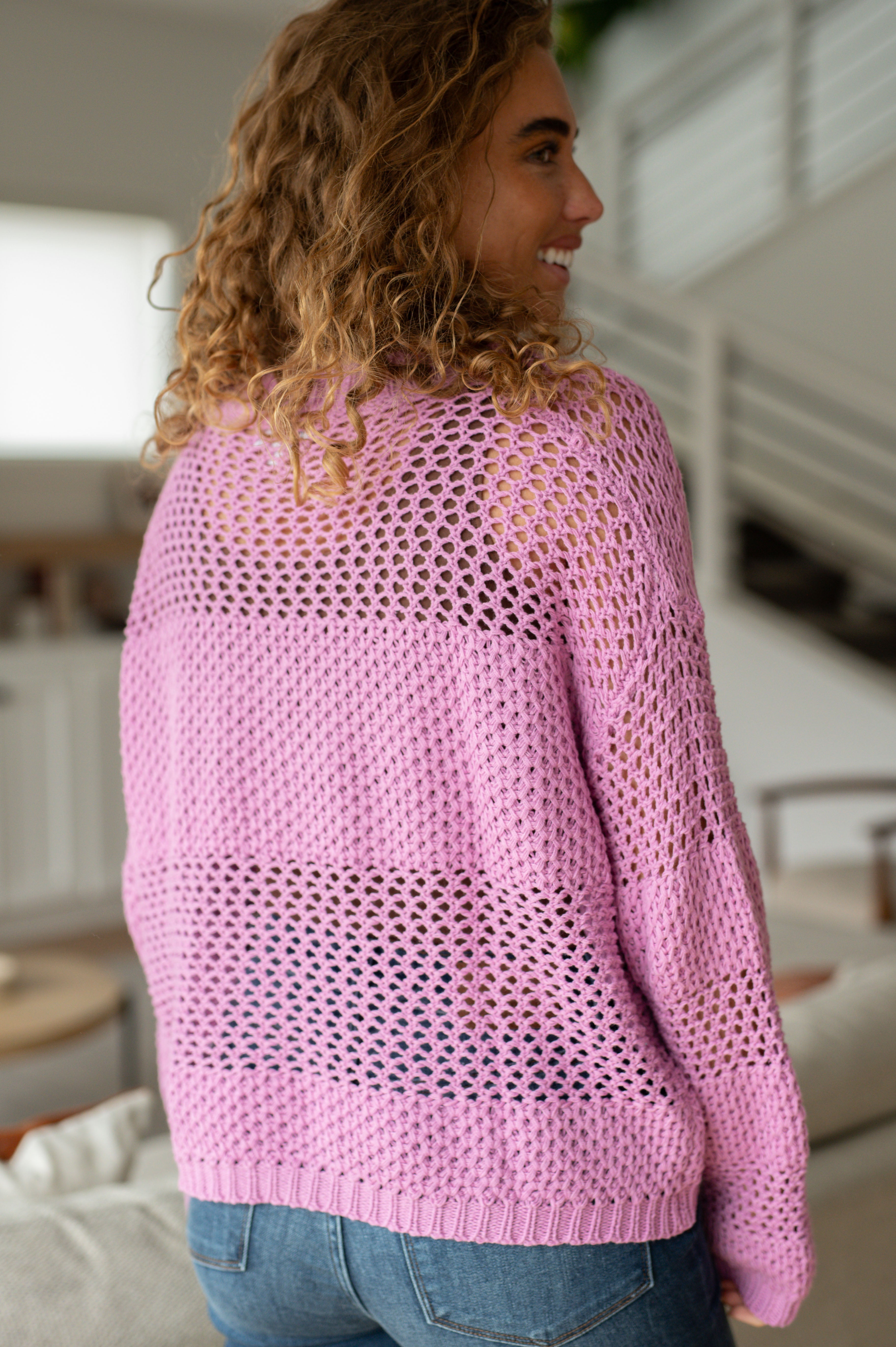 Woman with curly hair wearing a loose pink knit sweater with open weave detail standing indoors facing sideways