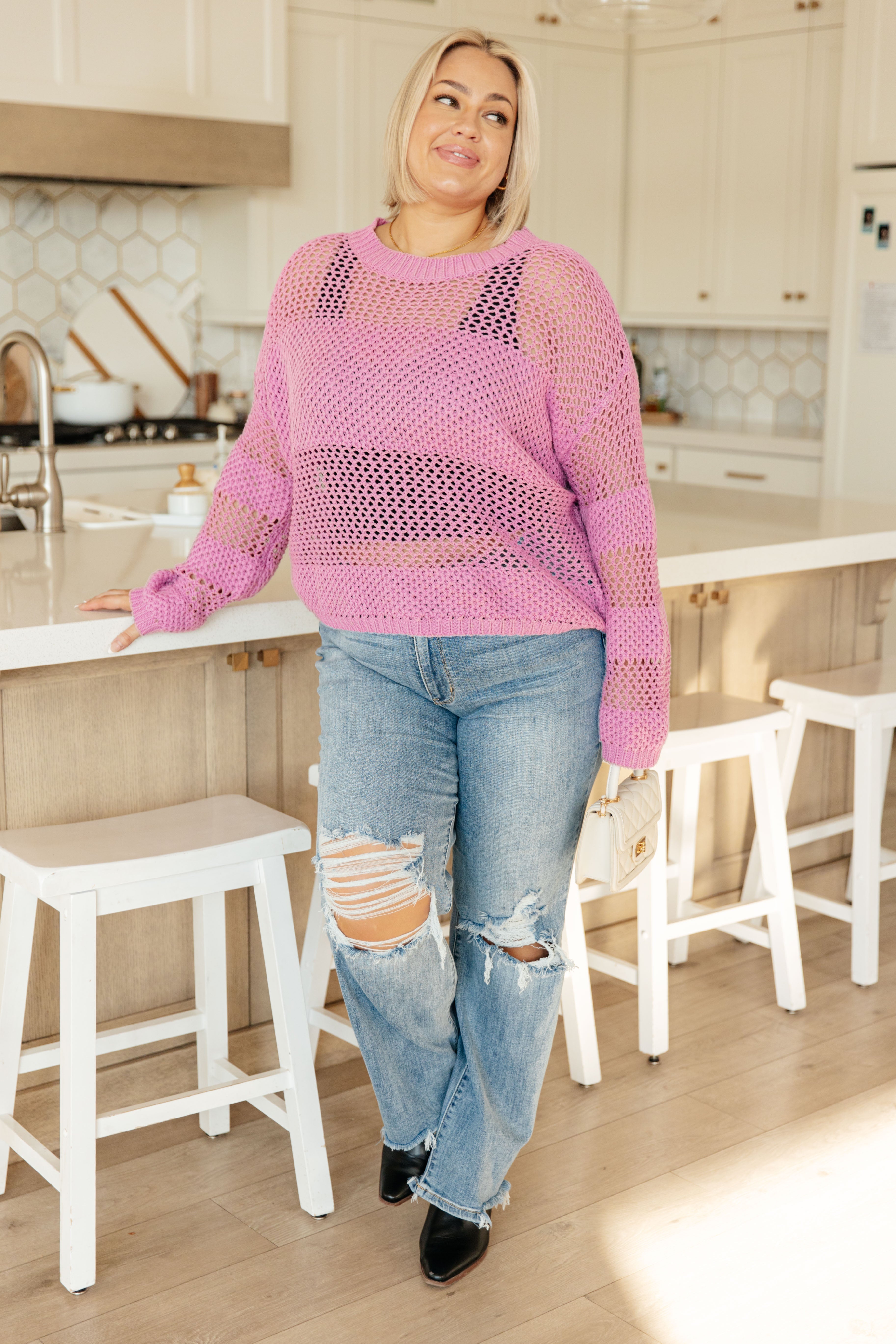 Plus-size woman wearing a loose pink open-knit sweater layered over a black crop top with ripped blue jeans indoors